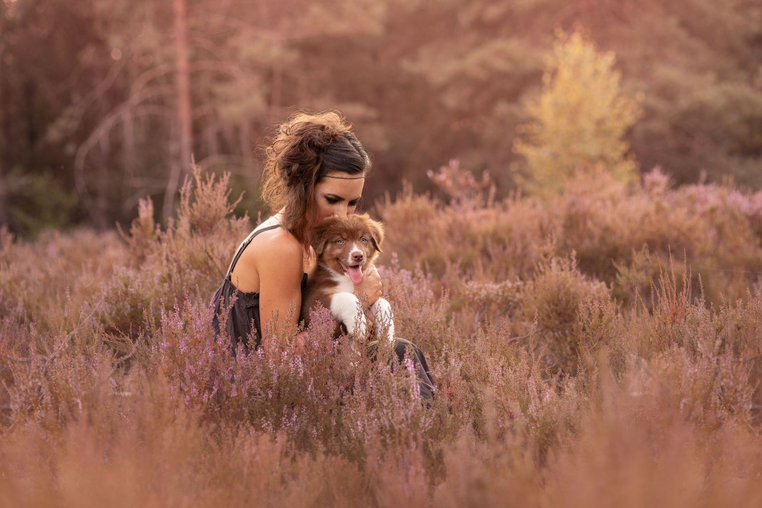 A young woman with curly hair tenderly embraces a small puppy while sitting in a field of purple heather.