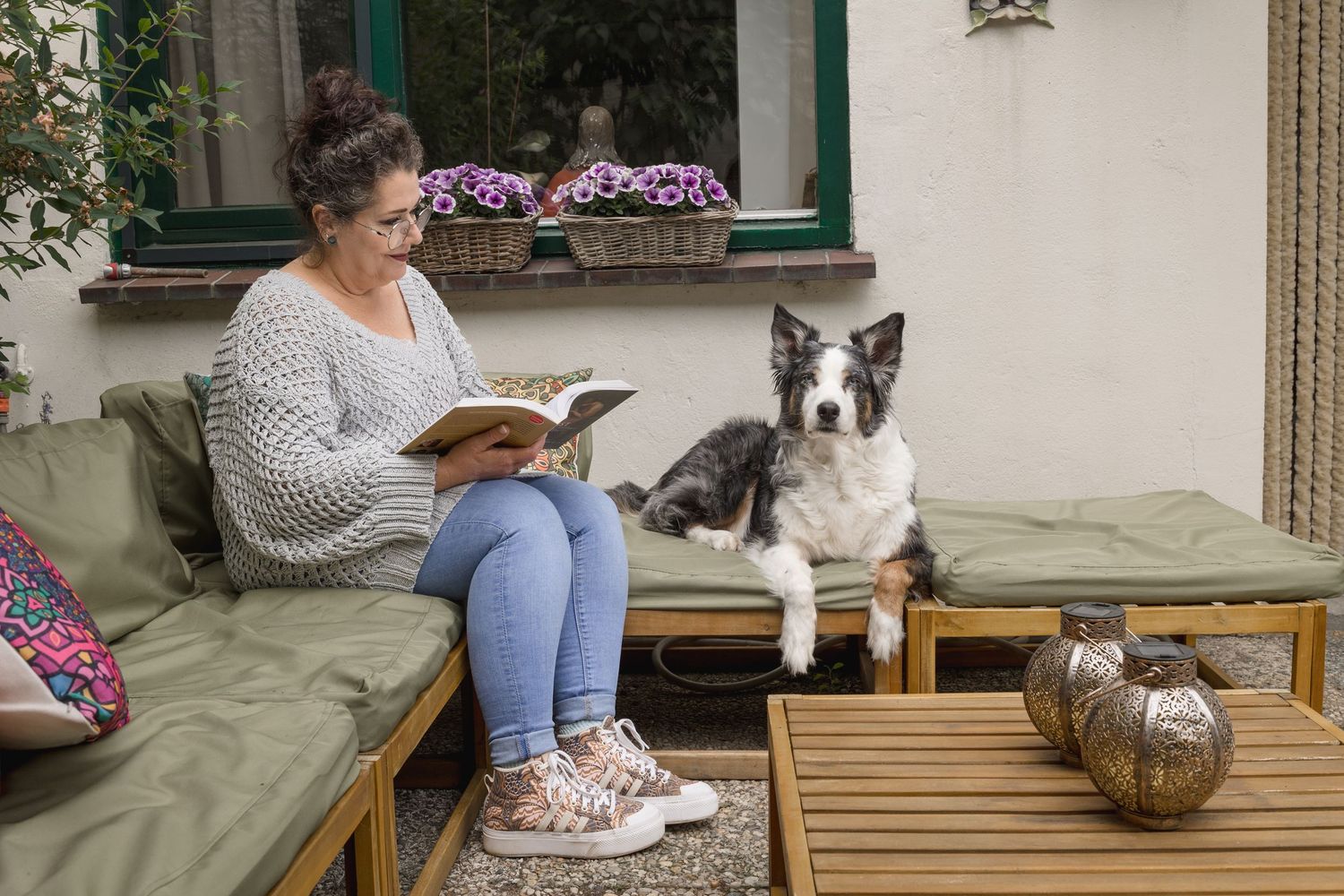 Woman in grey sweater reads book on outdoor patio sofa beside attentive Border Collie dog.