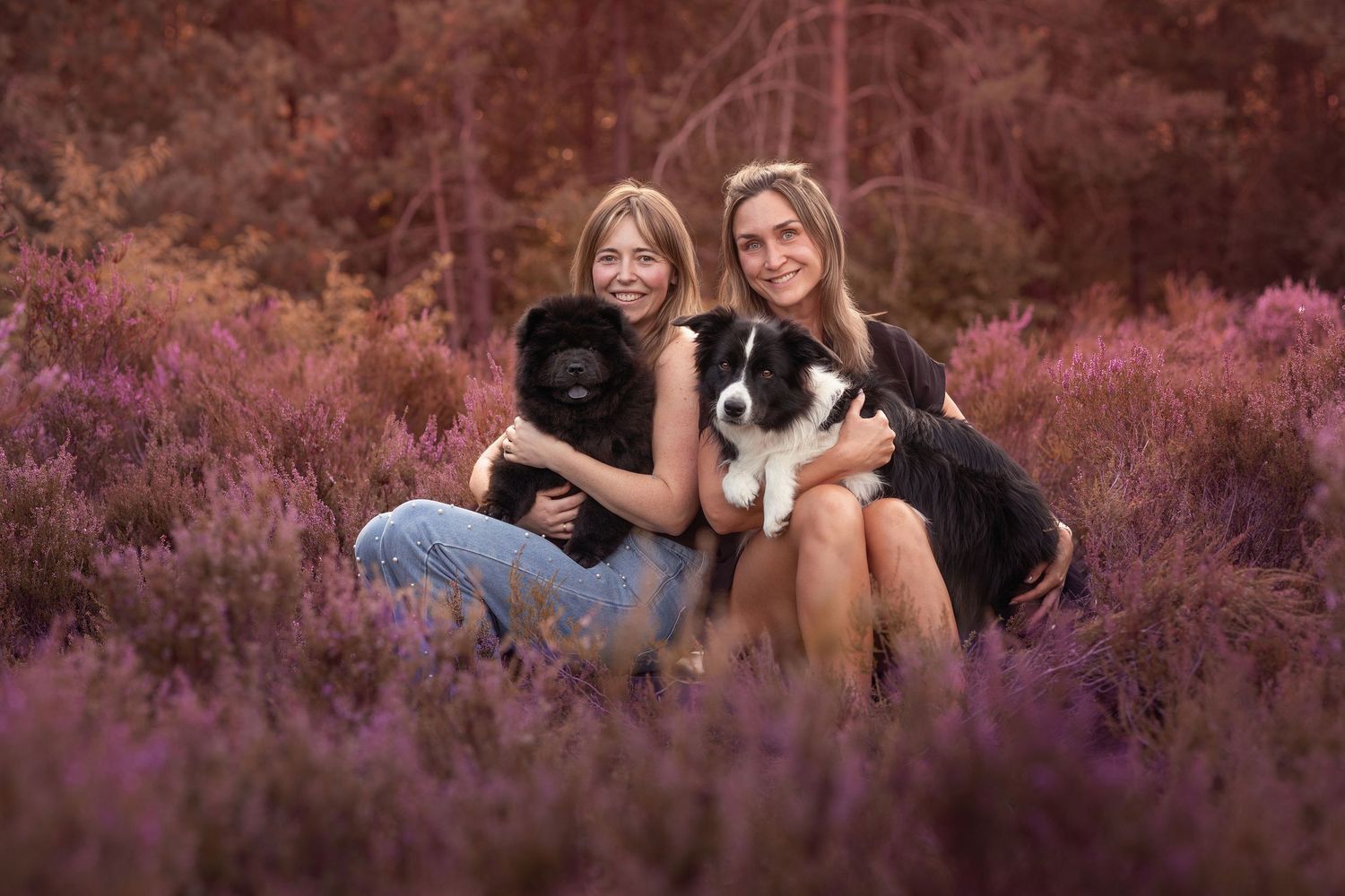 Two young women smile while holding a black Chow Chow and a Border Collie in a purple heather field.