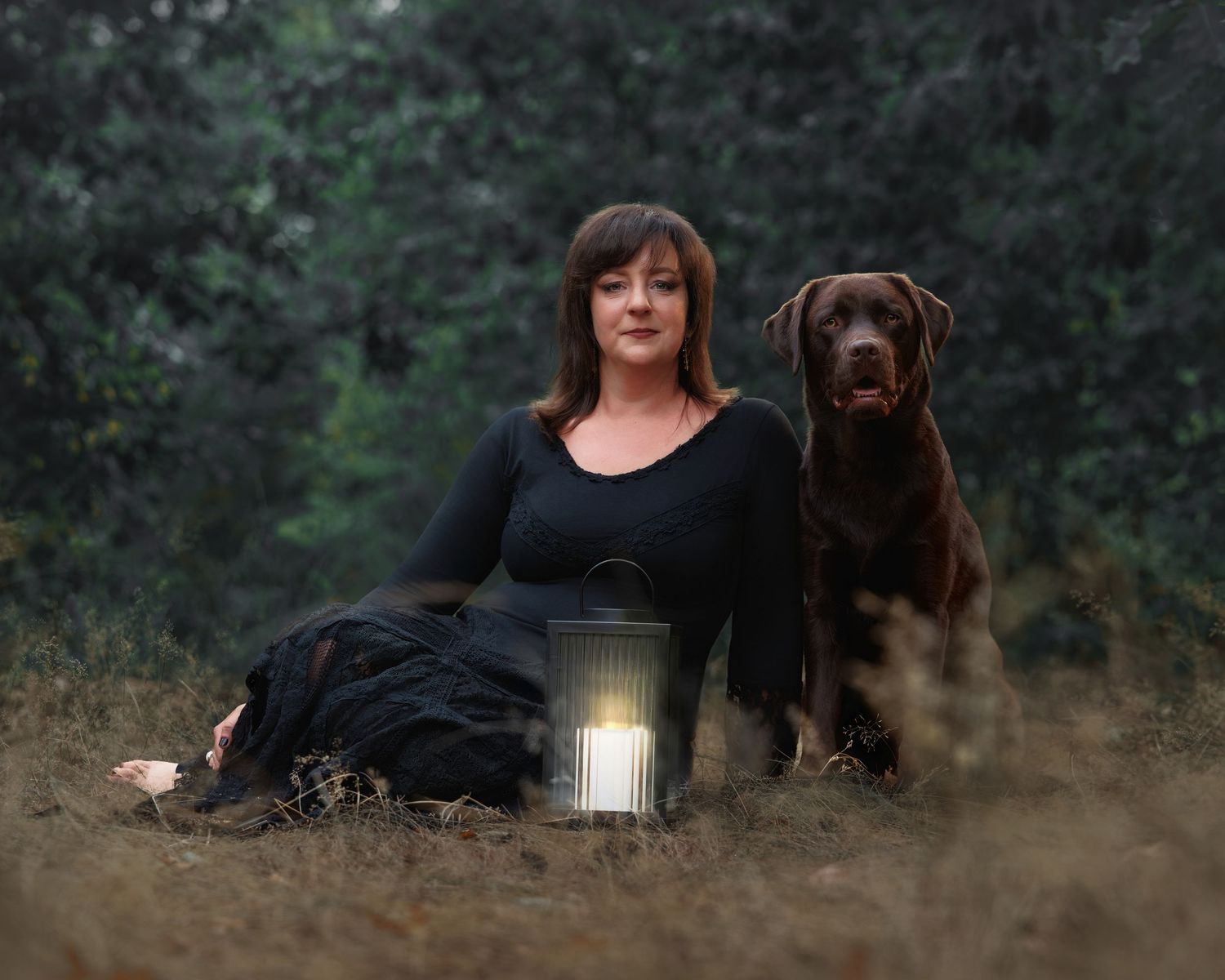 Brunette woman in black outfit sits beside a chocolate Labrador with a glowing lantern outdoors.