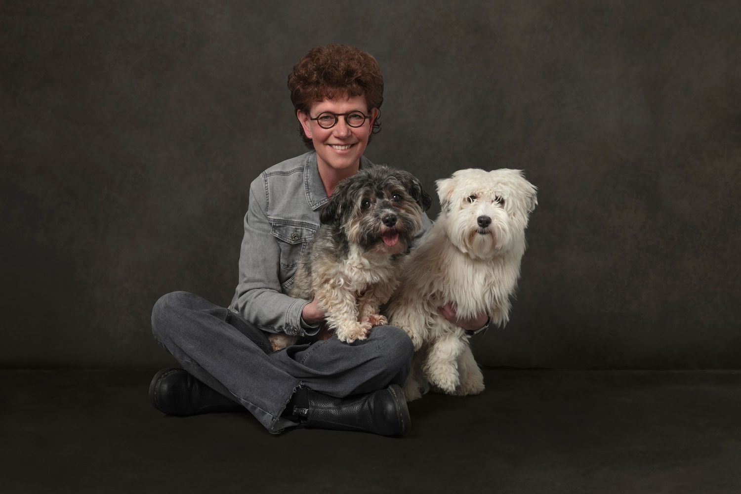 Person sitting cross-legged on floor smiling, holding two fluffy small dogs, photographed against a gray studio background.