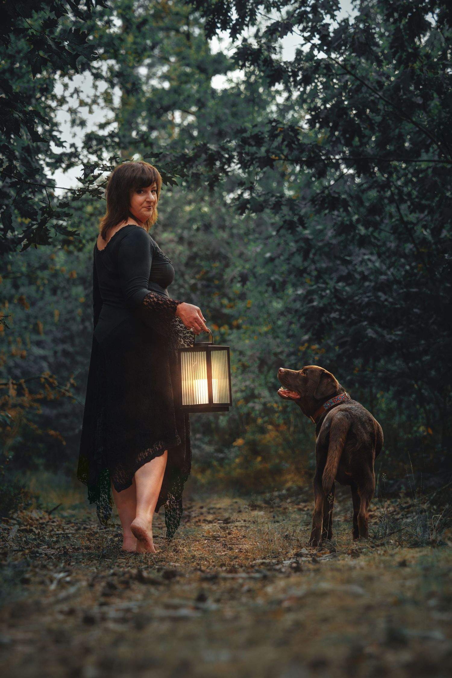 Woman in black dress holding glowing lantern stands barefoot in dark forest with brown Labrador dog at dusk.