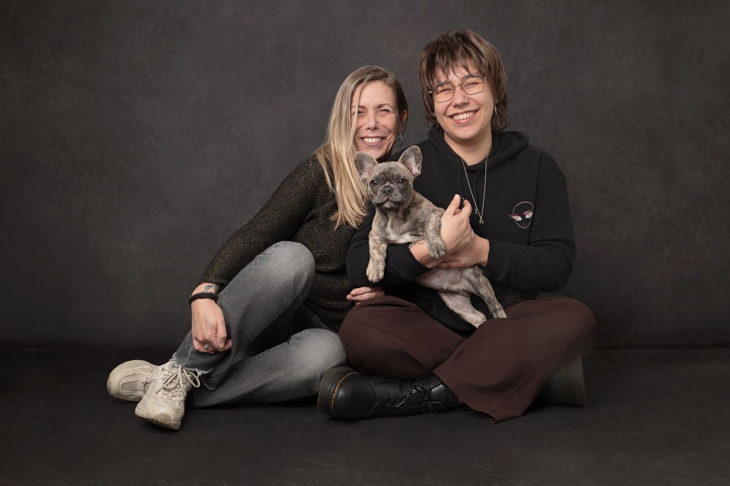 Two smiling women sit together on a studio floor holding a small grey puppy against a dark background.