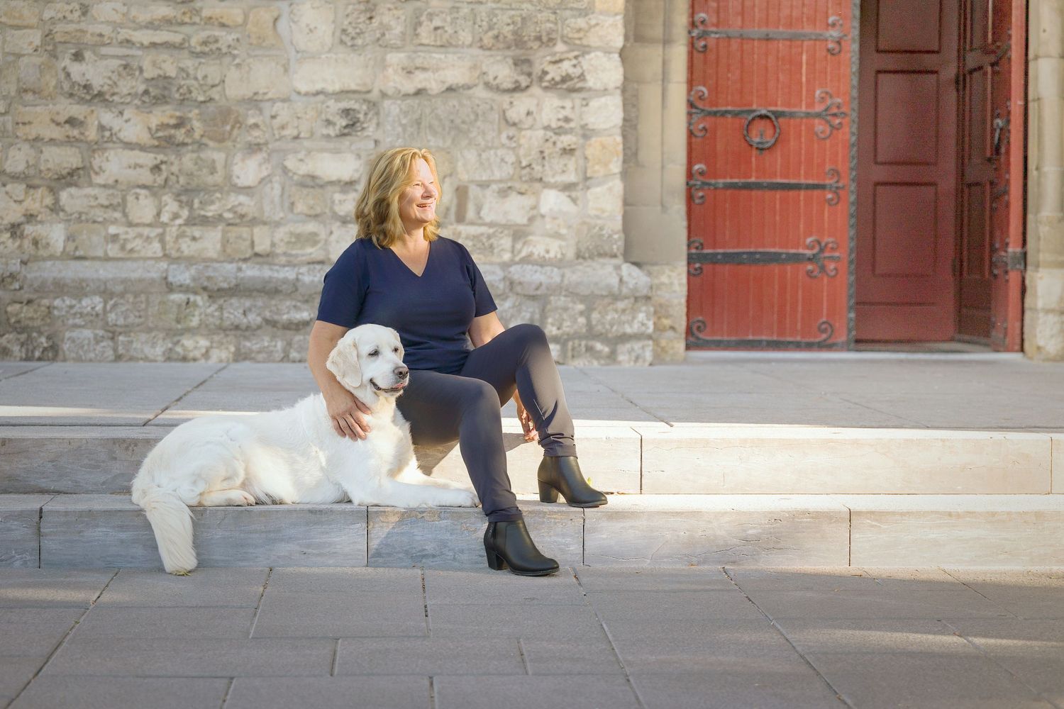 Woman sitting on stone steps with a white Labrador dog beside her, near a rustic red door and stone wall.