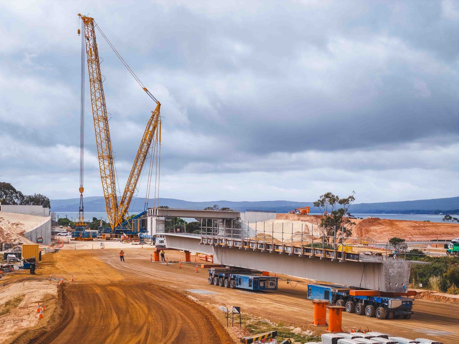 Workers and heavy cranes lift and position a long concrete bridge beam on multi-axle trailers at an active highway construction site under c