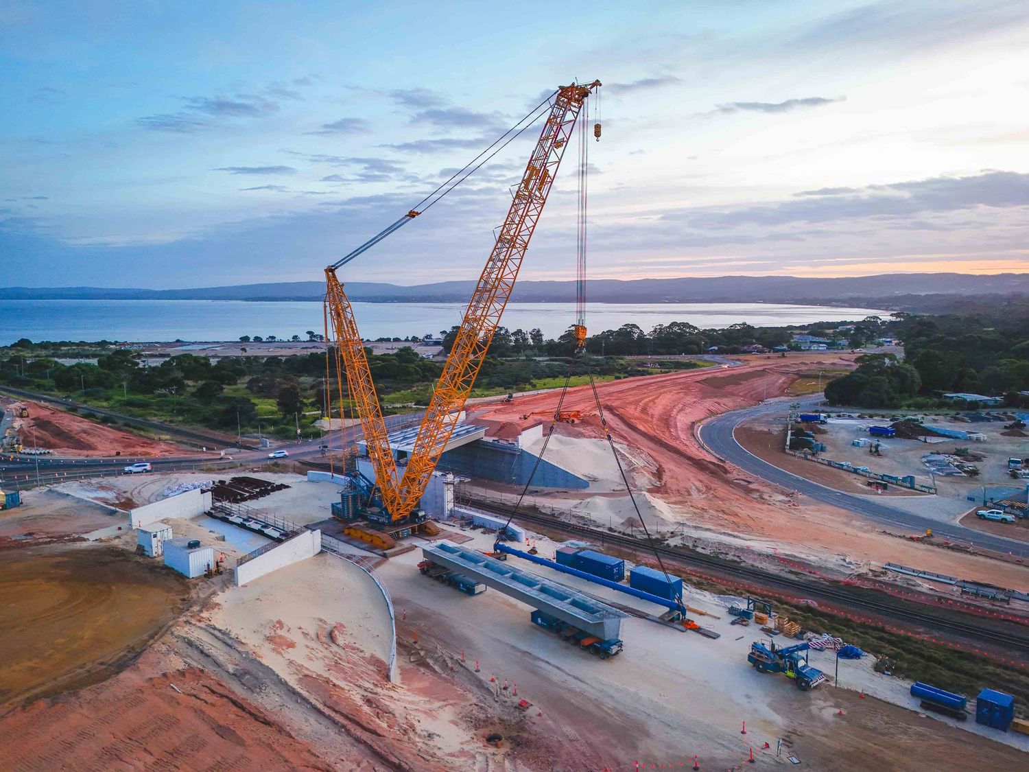 Aerial view of a large crane lifting concrete beams at a coastal road and bridge construction site at dusk, showing heavy machinery, earthwo