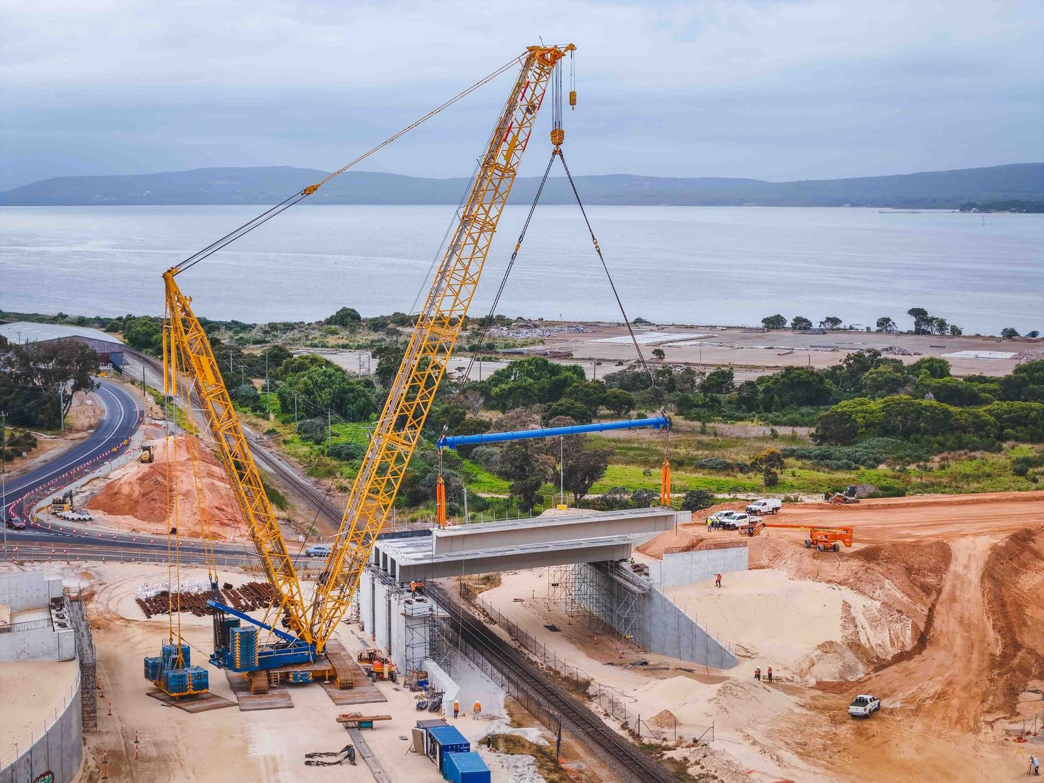 A large lattice boom crane lifts a concrete bridge beam into place at a coastal highway and railway construction site, with workers, vehicle