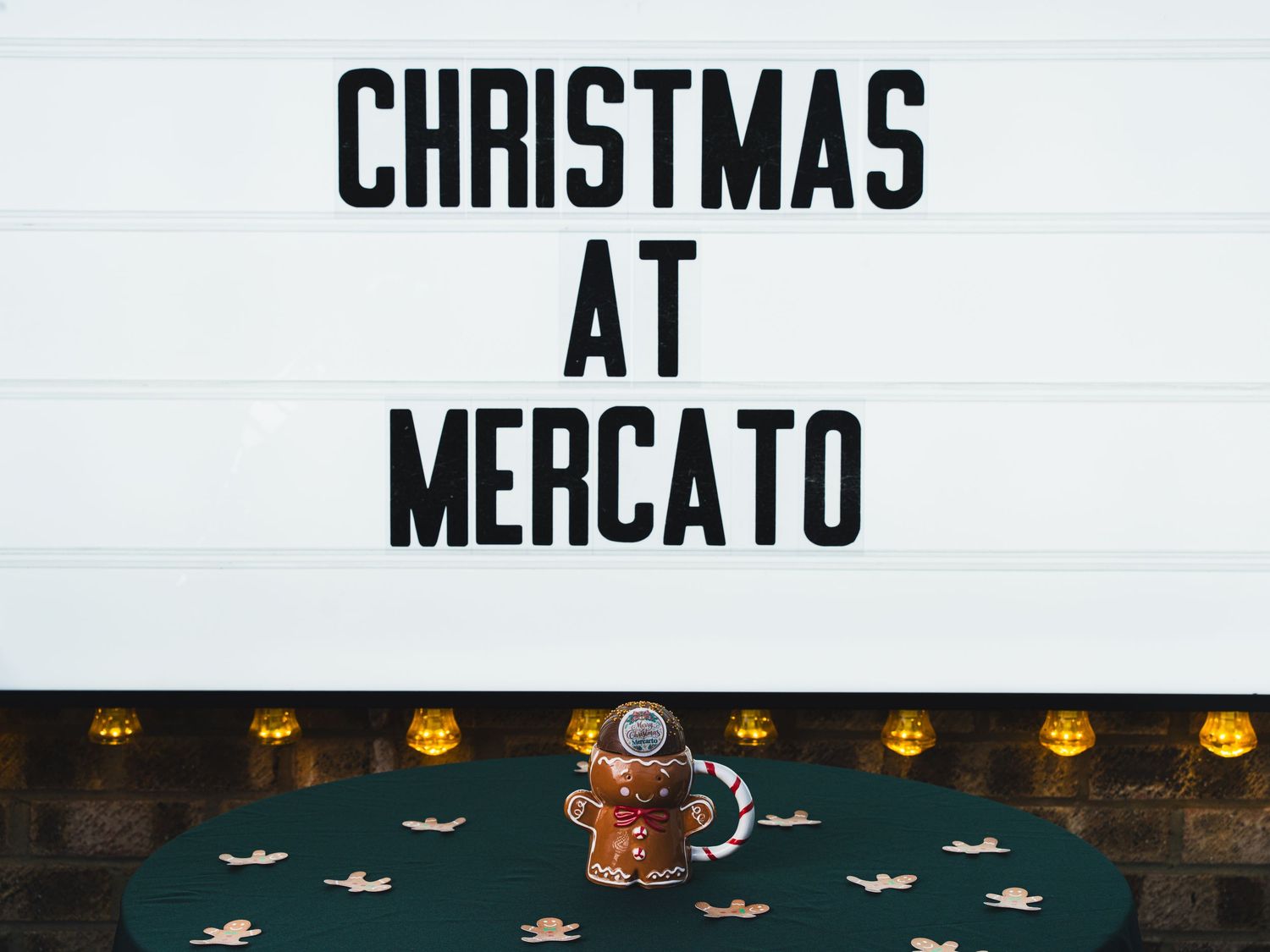 A marquee sign reading Christmas at Mercato above a green table with a gingerbread man mug and small snowflake decorations.