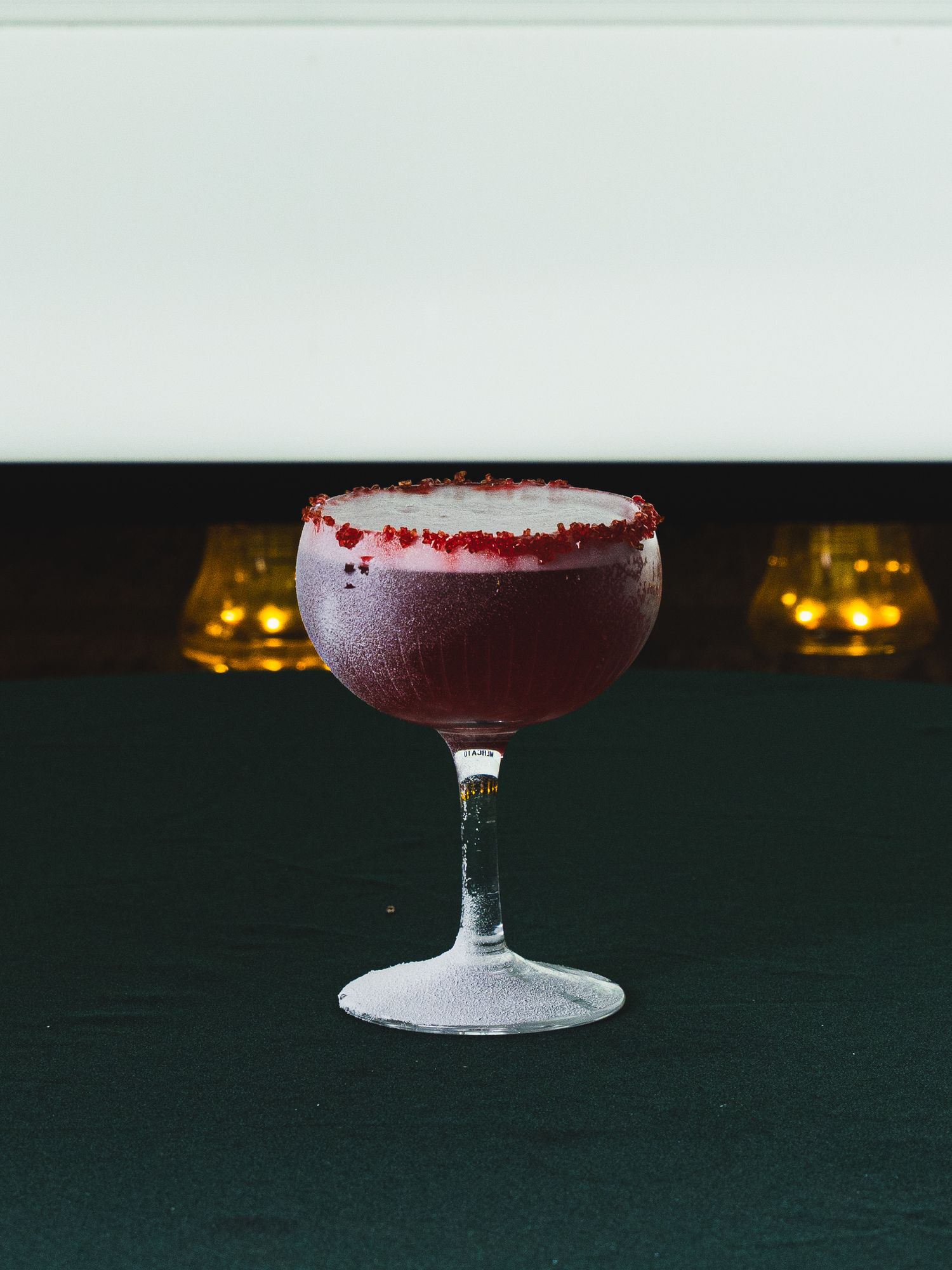 Deep red cocktail with a frothy foam top and sugared red rim, served in a vintage coupe glass on a dark table with candles behind.