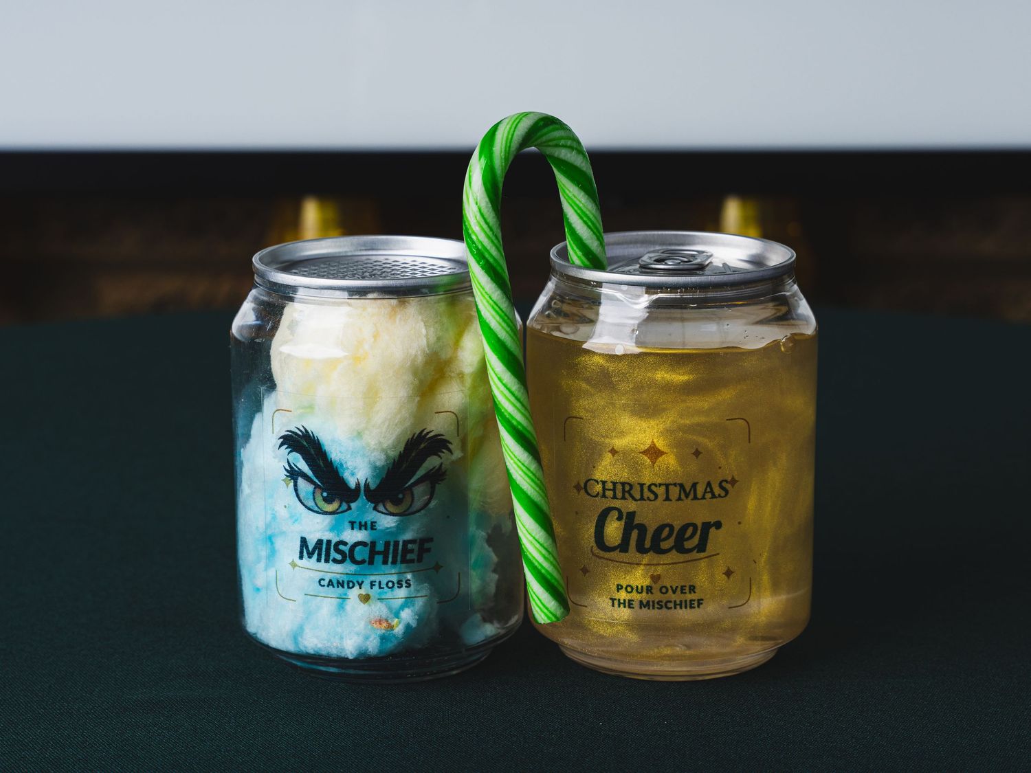 Two festive canned cocktails, The Mischief with blue cotton candy and Christmas Cheer with golden drink, joined by a green candy cane.