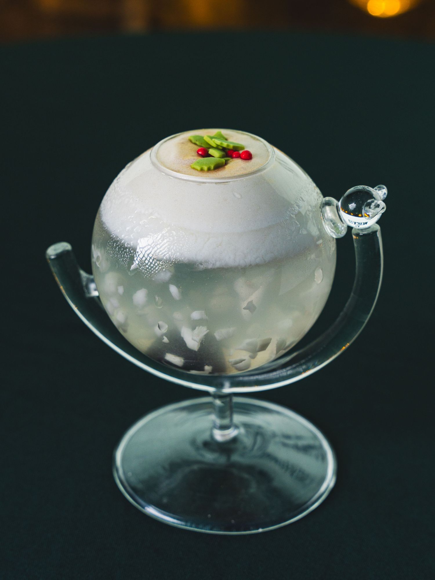 Festive cocktail served in a globe-shaped glass on a curved stand, topped with frothy foam and a holly leaf and red berry garnish.