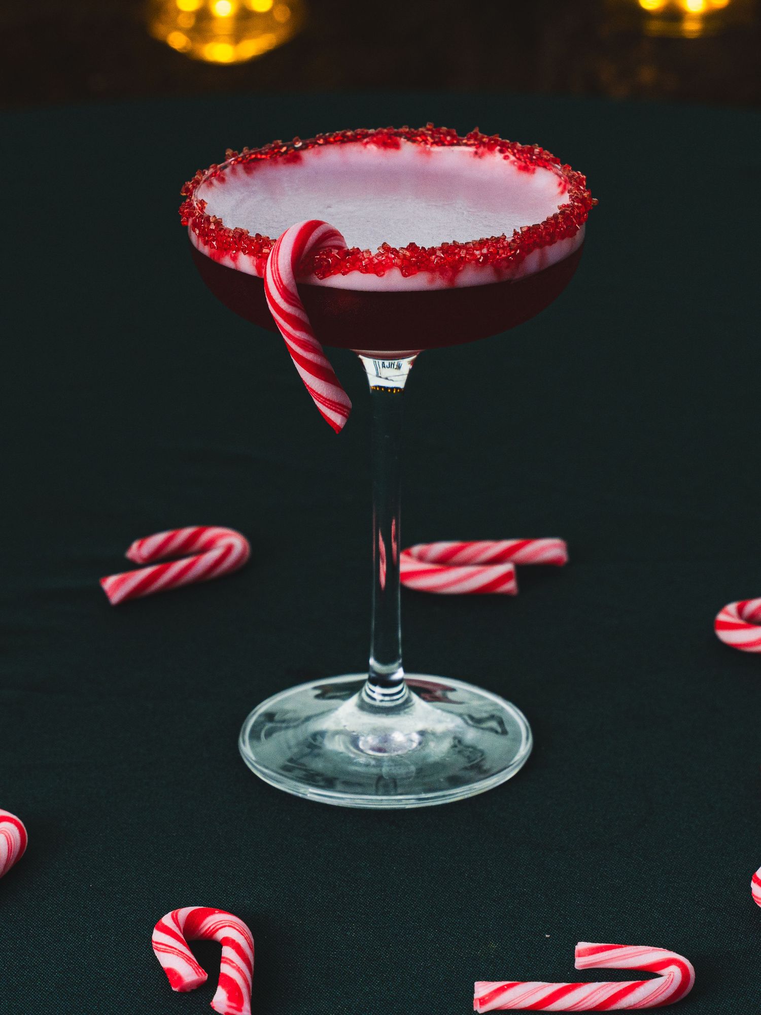 Festive peppermint cocktail in a coupe glass with a red sugar rim and candy cane garnish, surrounded by candy canes on a dark table.