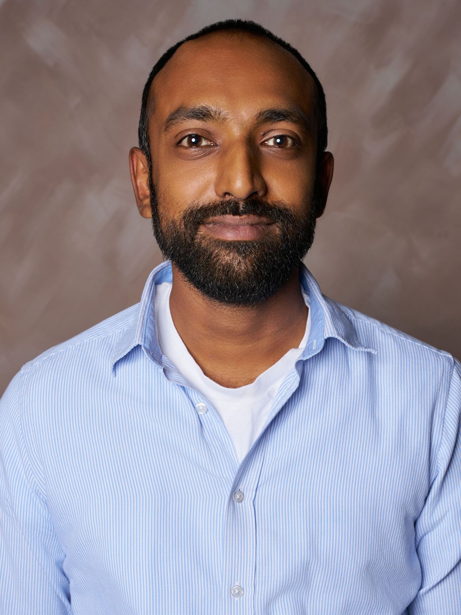Smiling man with a trimmed beard wearing a light blue striped button-up shirt over a white tee against a textured brown backdrop.