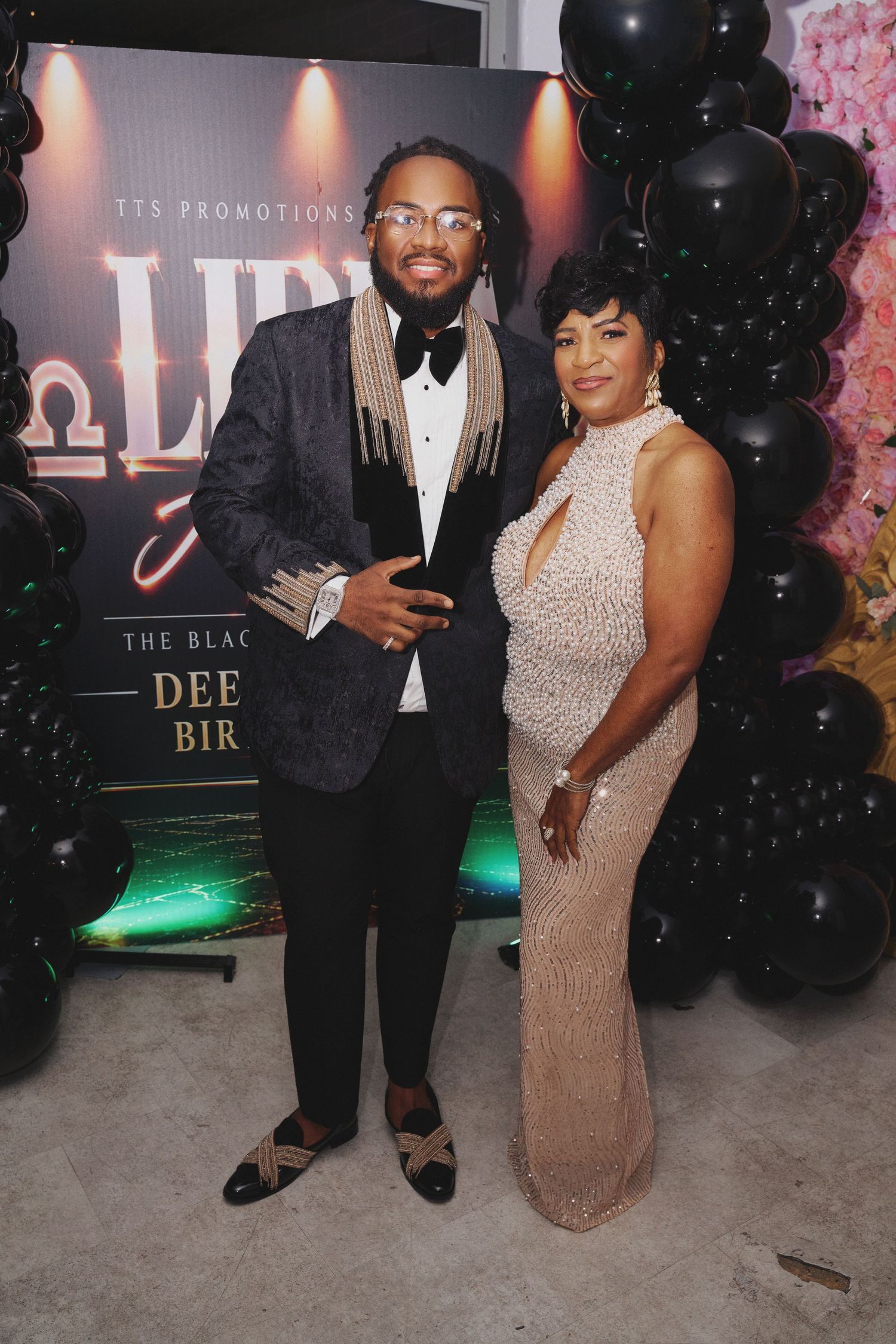 A man in a black tuxedo jacket and bow tie poses with a woman in a beaded champagne gown at a black-balloon birthday event.