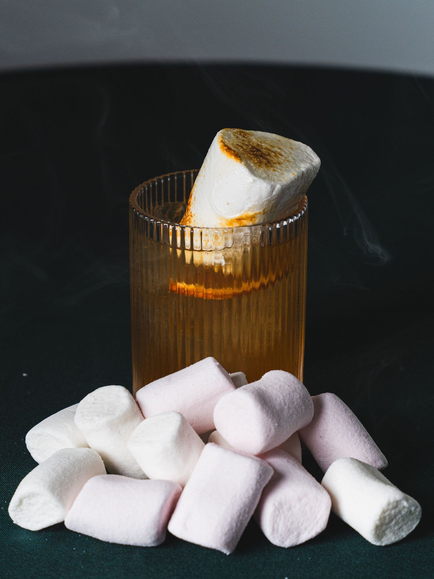A fluted glass of amber cocktail topped with a toasted marshmallow, surrounded by pink and white marshmallows on a dark green surface.