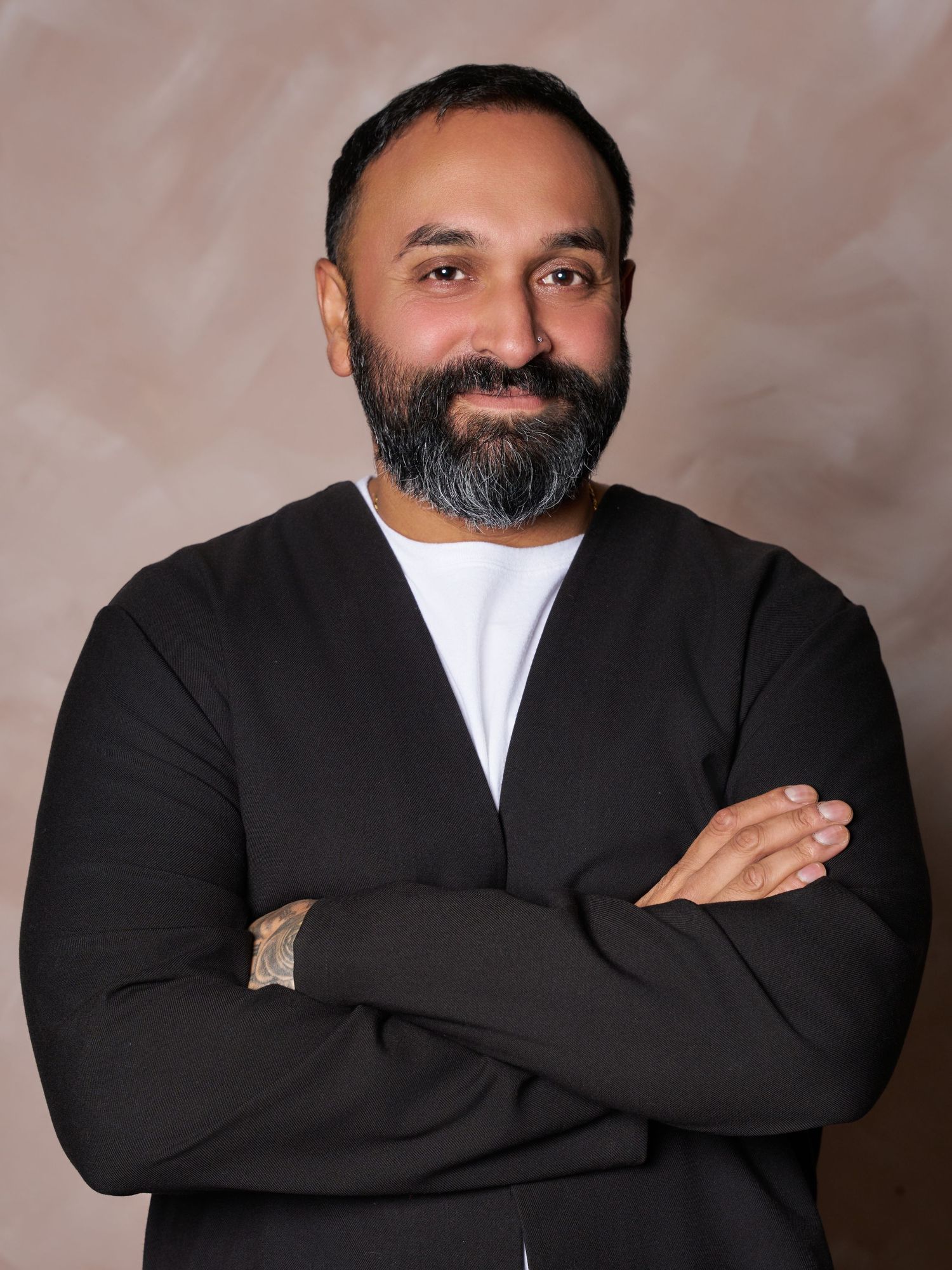 Smiling bearded man in a black cardigan and white t-shirt posing with arms crossed against a warm beige textured studio backdrop.