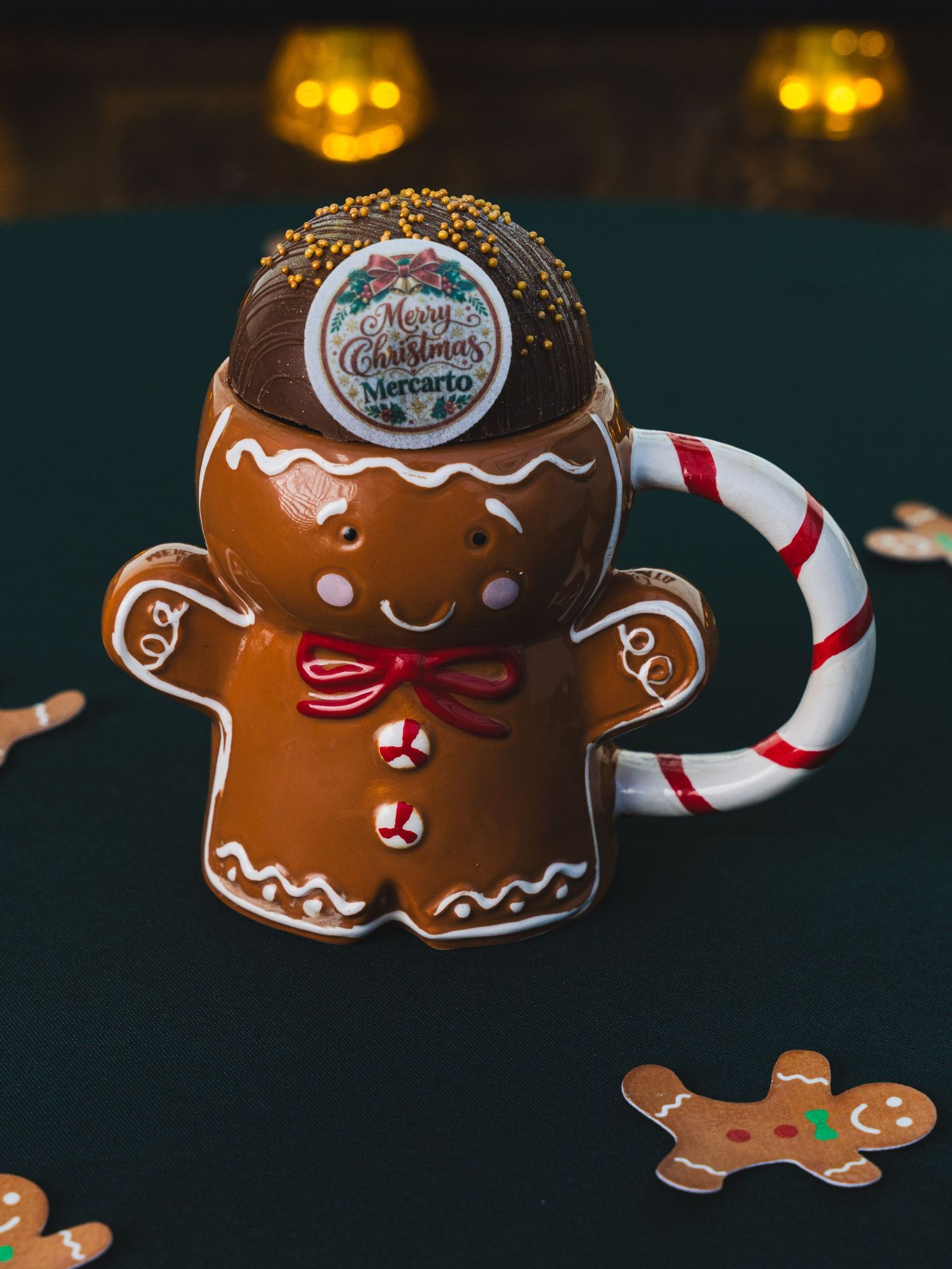 Gingerbread man ceramic mug with candy cane handle, topped with a Mercarto Merry Christmas hot chocolate bomb on a dark festive table.