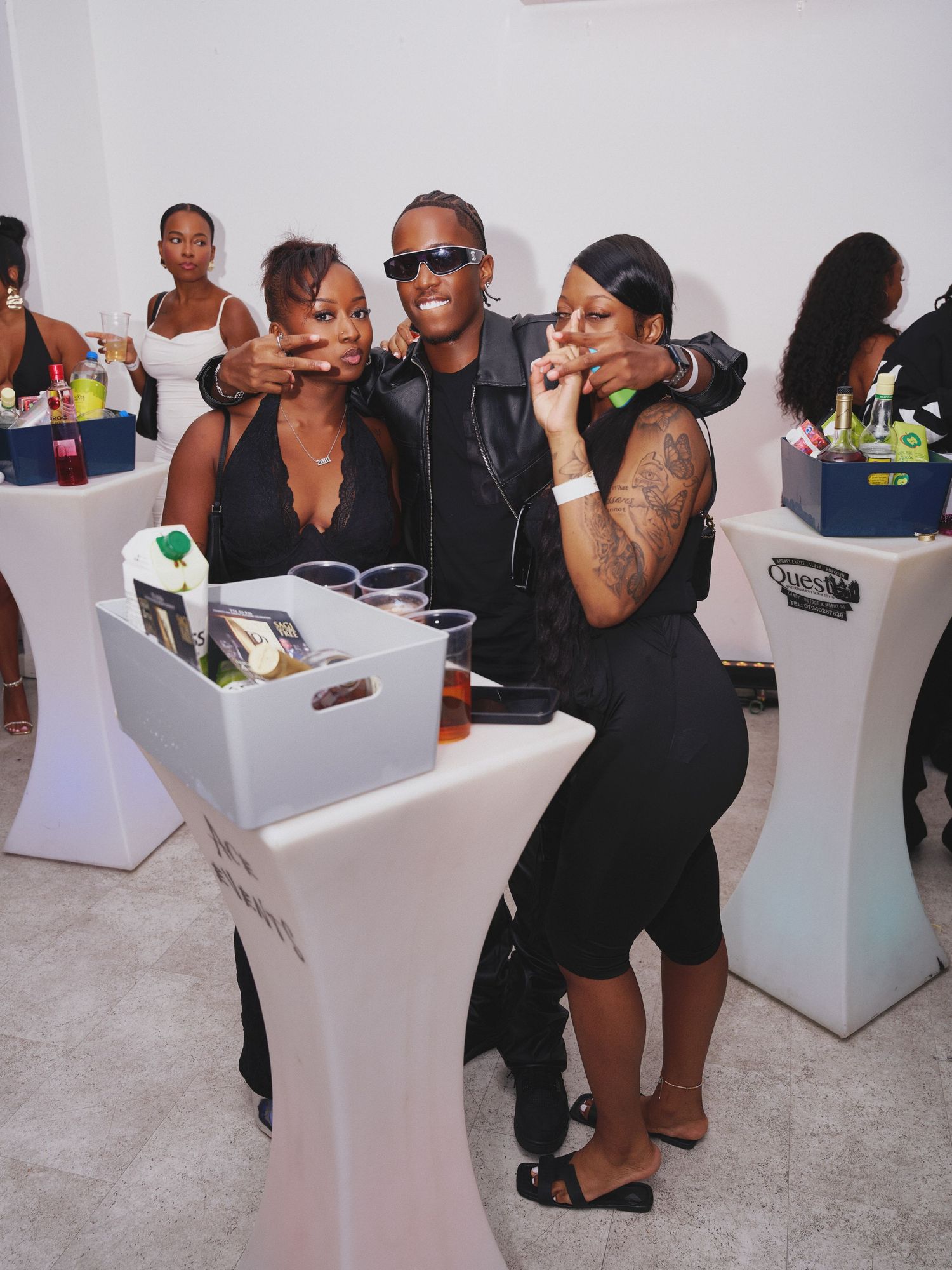 Three friends in black outfits pose and gesture together at a lively party beside white cocktail tables with drinks and bottles.