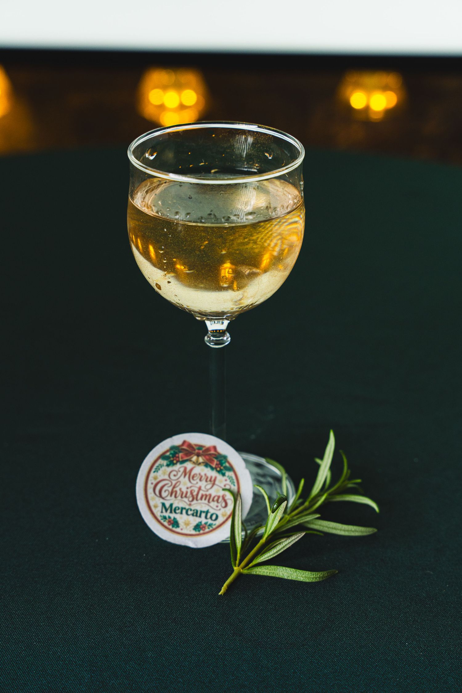 Glass of white wine on a dark green table with a rosemary sprig and a Merry Christmas Mercarto sticker.