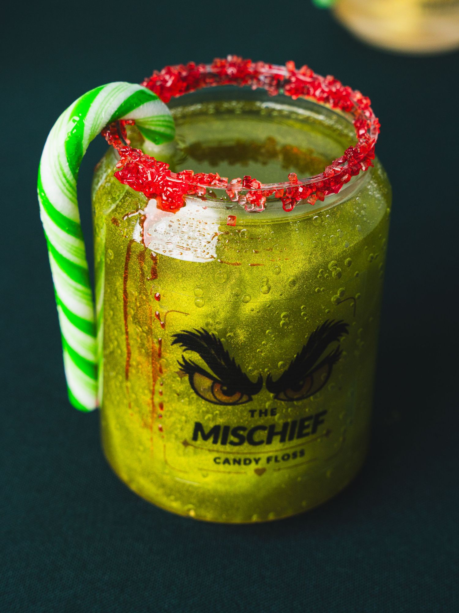 Green Grinch-inspired cocktail in a jar with a red sugar-rimmed edge, striped candy cane garnish and The Mischief Candy Floss label.