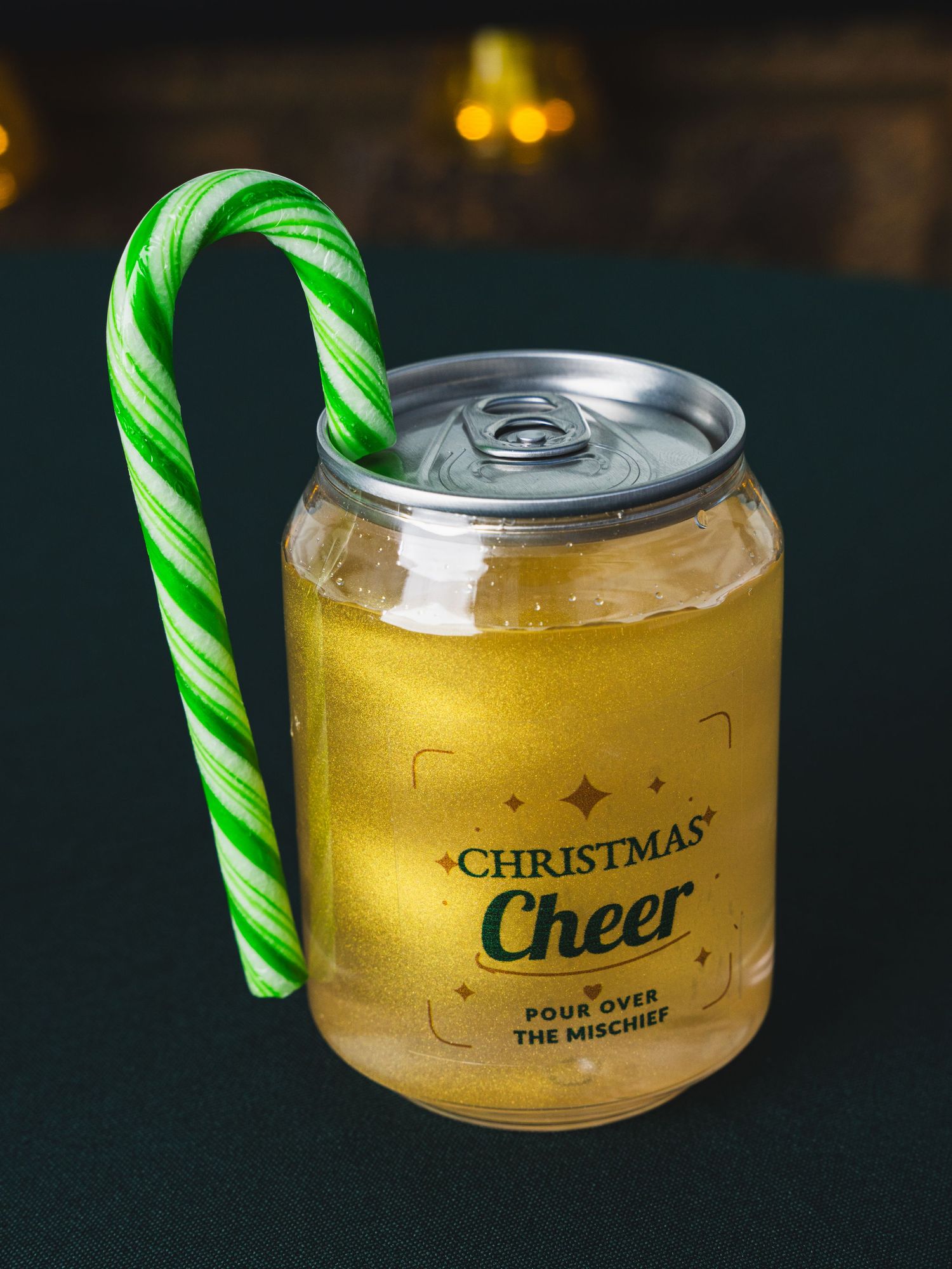 Can-shaped glass filled with shimmering gold drink, labeled Christmas Cheer, with a green striped candy cane hooked on the rim.