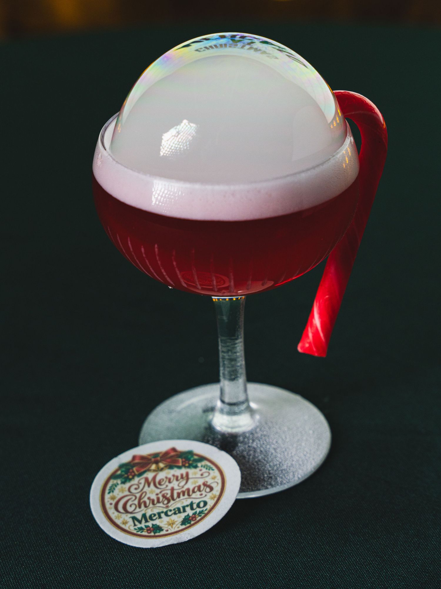 Red Christmas cocktail topped with a glossy bubble dome and pink foam in a stemmed glass, with a Merry Christmas Mercarto coaster.