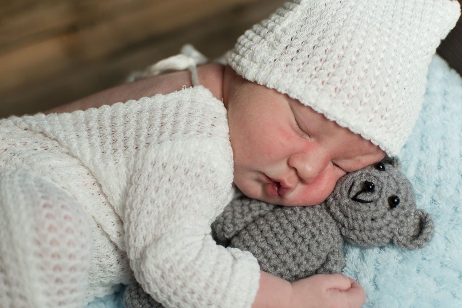 Sleeping infant in white knitted outfit snuggles with gray stuffed animal on light blue blanket.