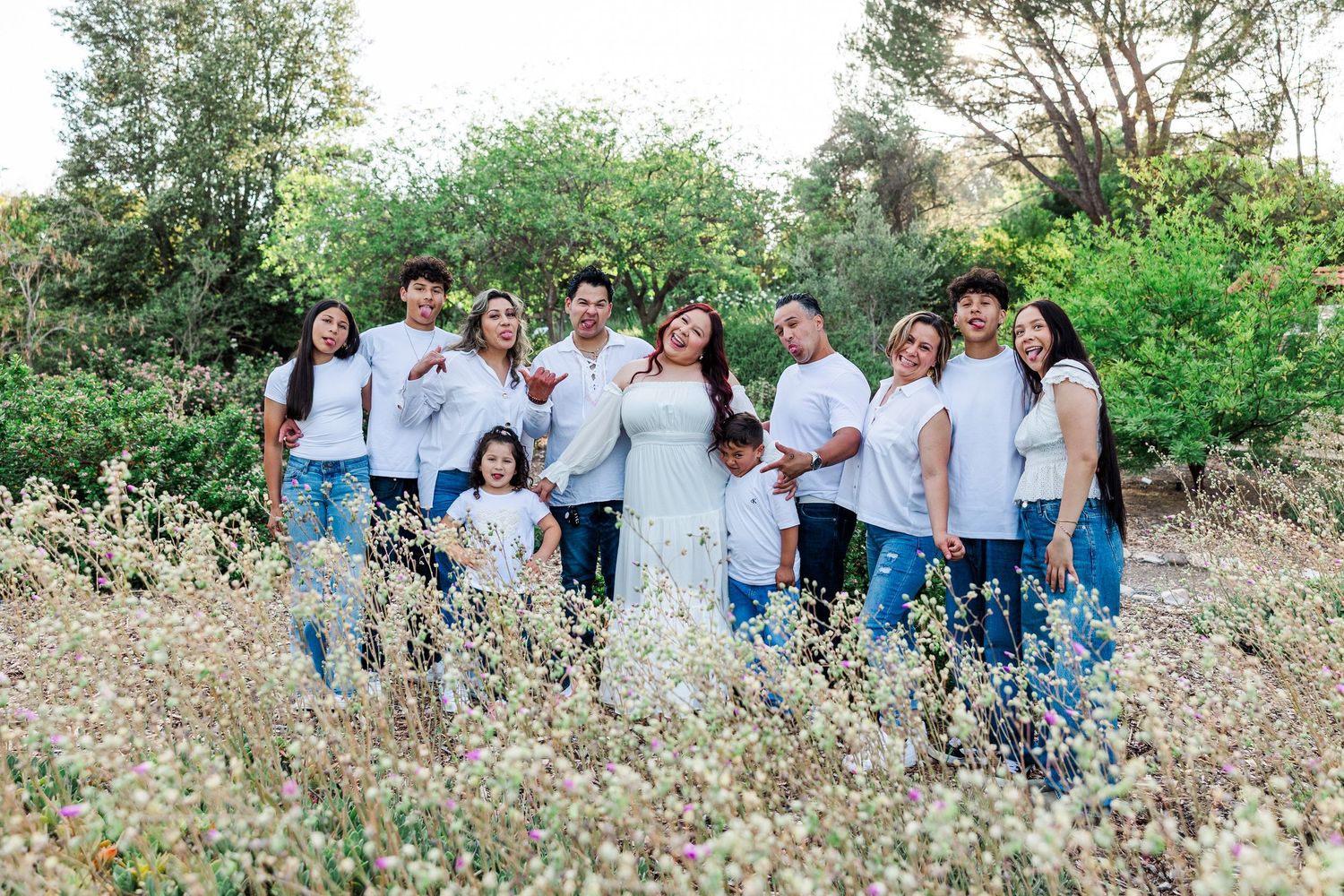 A group wearing white and blue casual attire poses together outdoors among flowering meadow grass and trees.