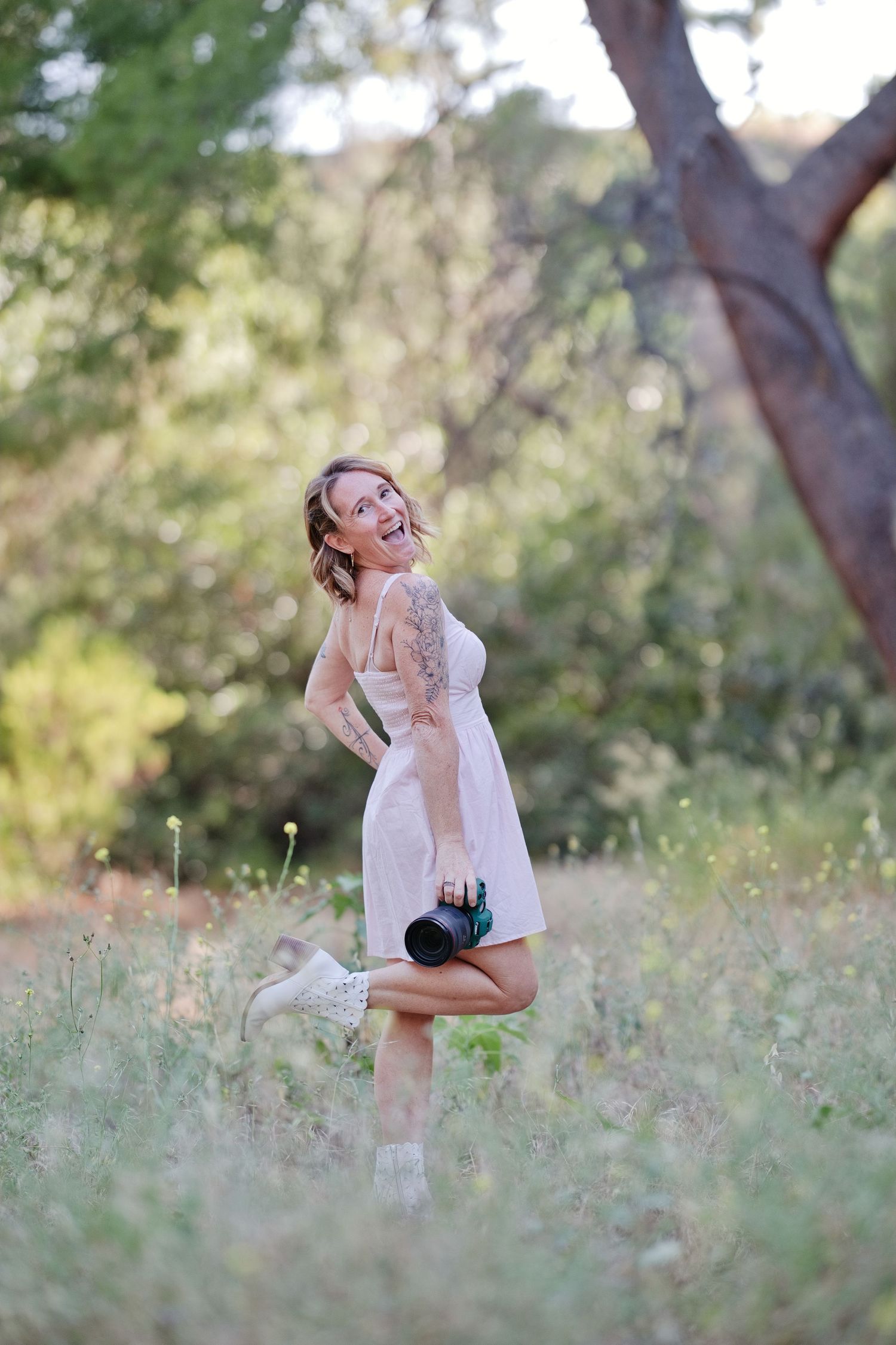 Woman in pink dress holding camera smiling in a sunlit forest meadow with tattoos visible on her arm.