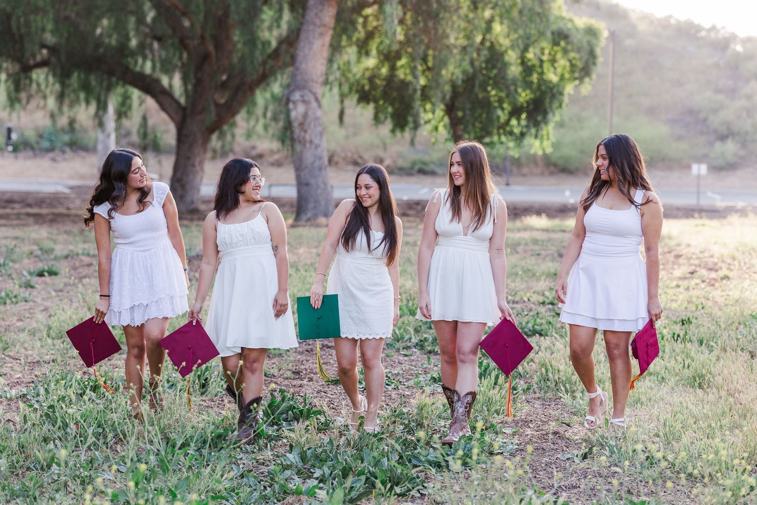 A group of people walk through a sun-lit grassy field carrying burgundy bags while wearing white outfits.