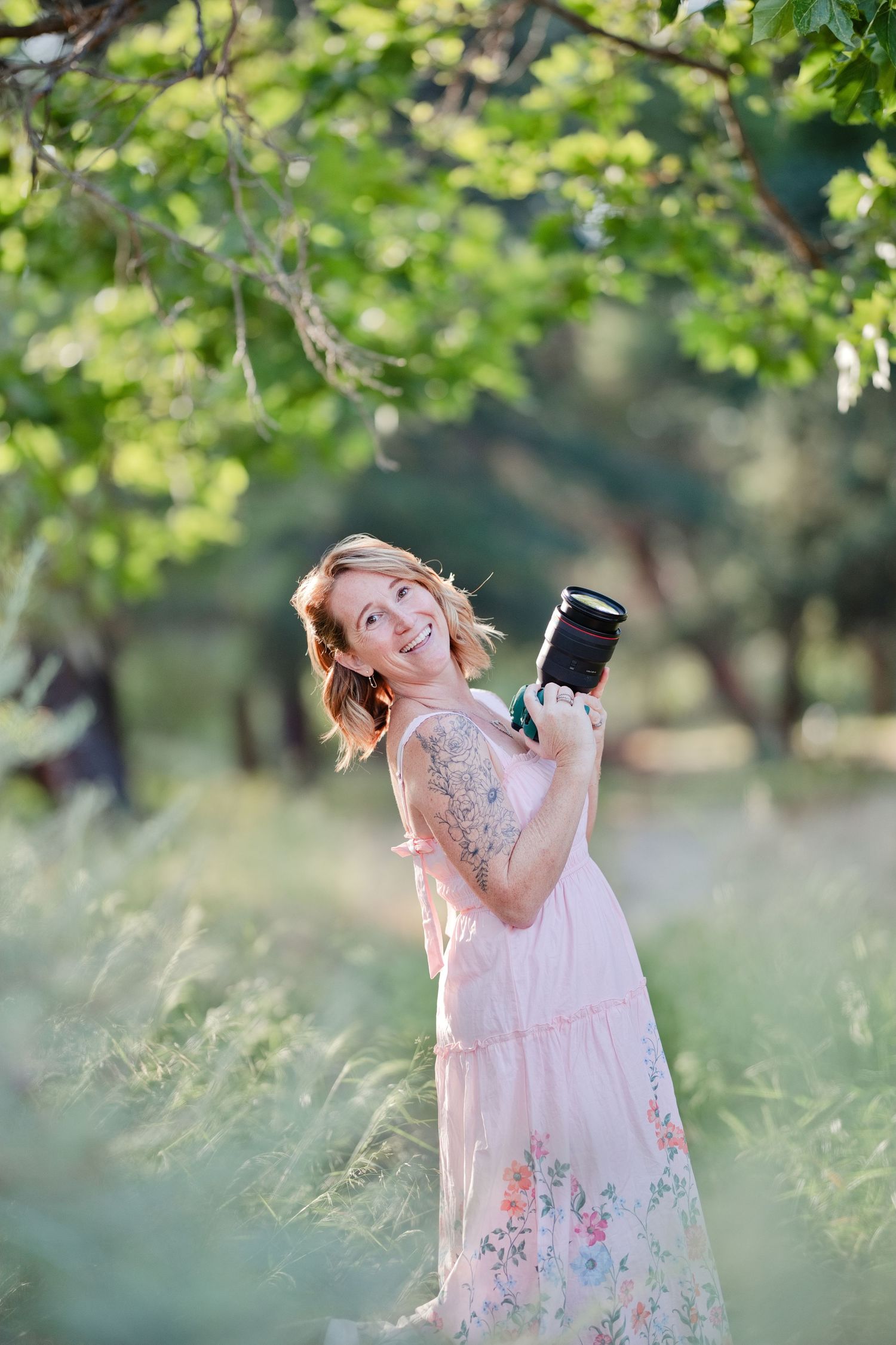 Smiling woman in pink floral dress holding a camera outdoors in a lush green garden setting.