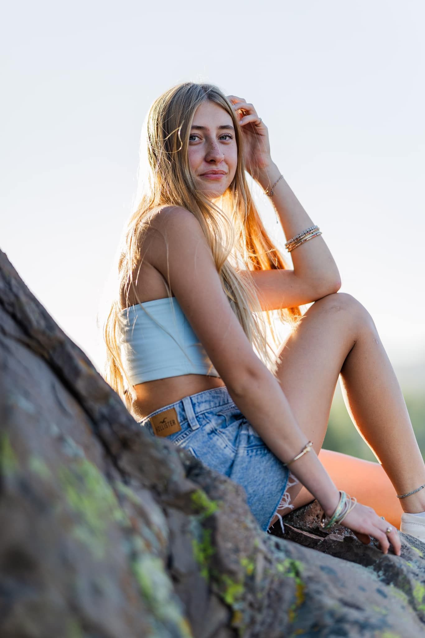 Sun sets behind someone wearing a white crop top and denim shorts sitting on mossy rocks during golden hour.
