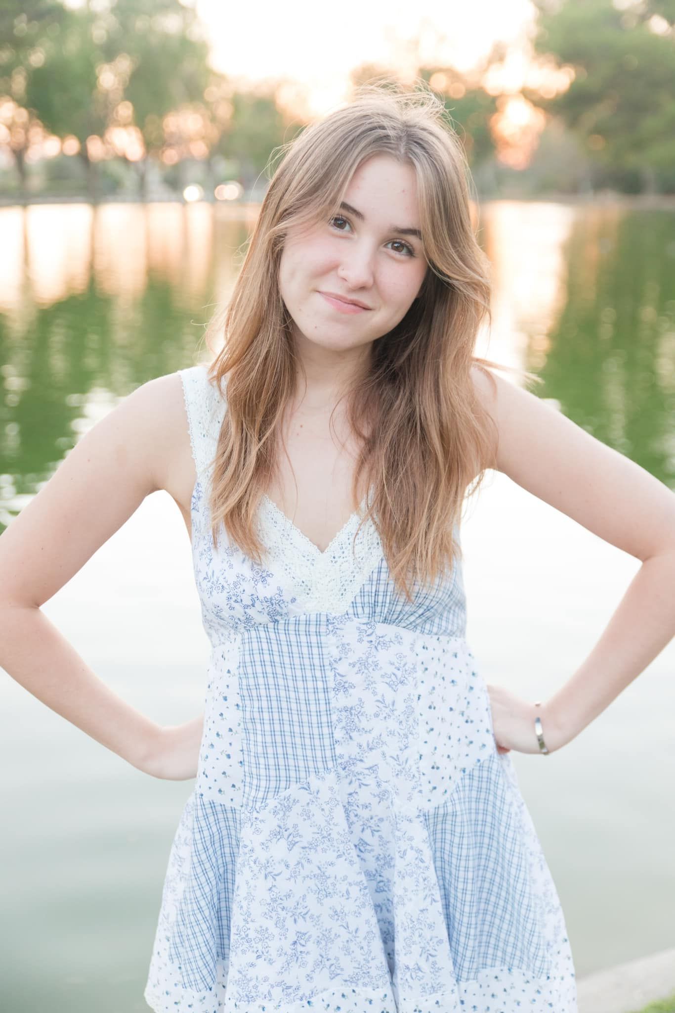 A person in a light blue and white gingham sundress poses outdoors during golden hour near a reflective lake.