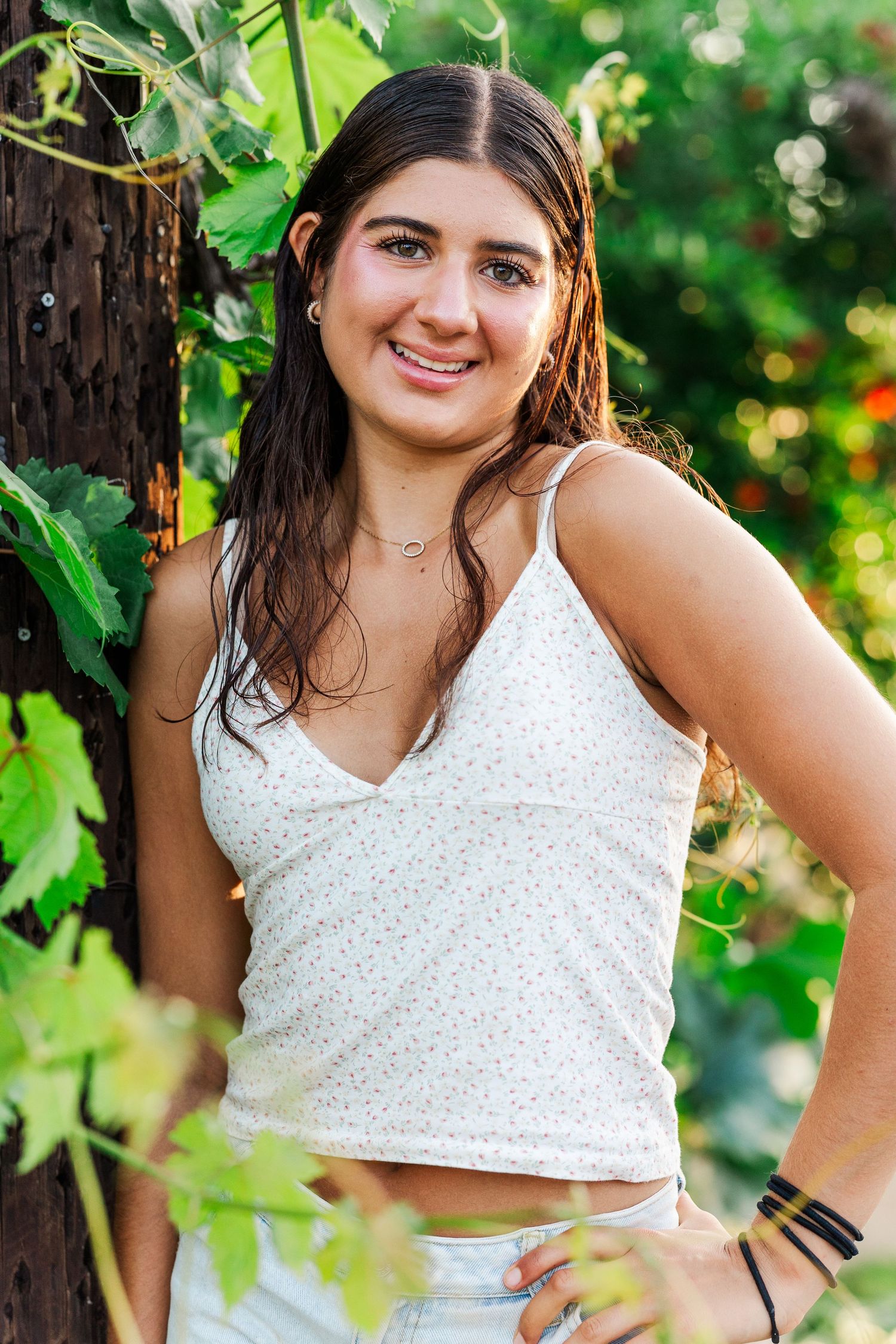 A person in a white sleeveless top poses outdoors among green foliage in a summer garden setting.