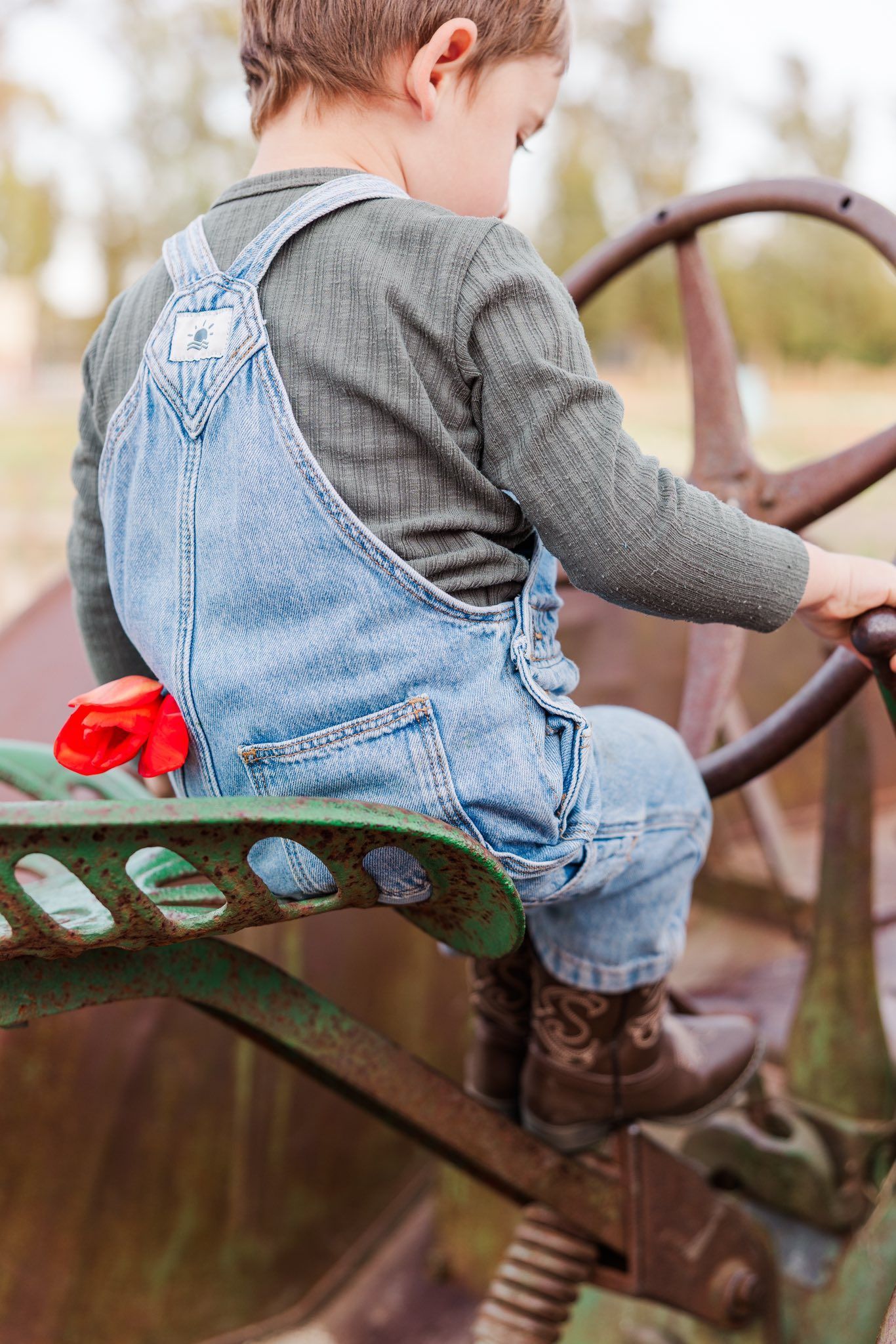 Little one in denim overalls and gray sweater climbing on vintage tractor seat with red bandana in pocket.