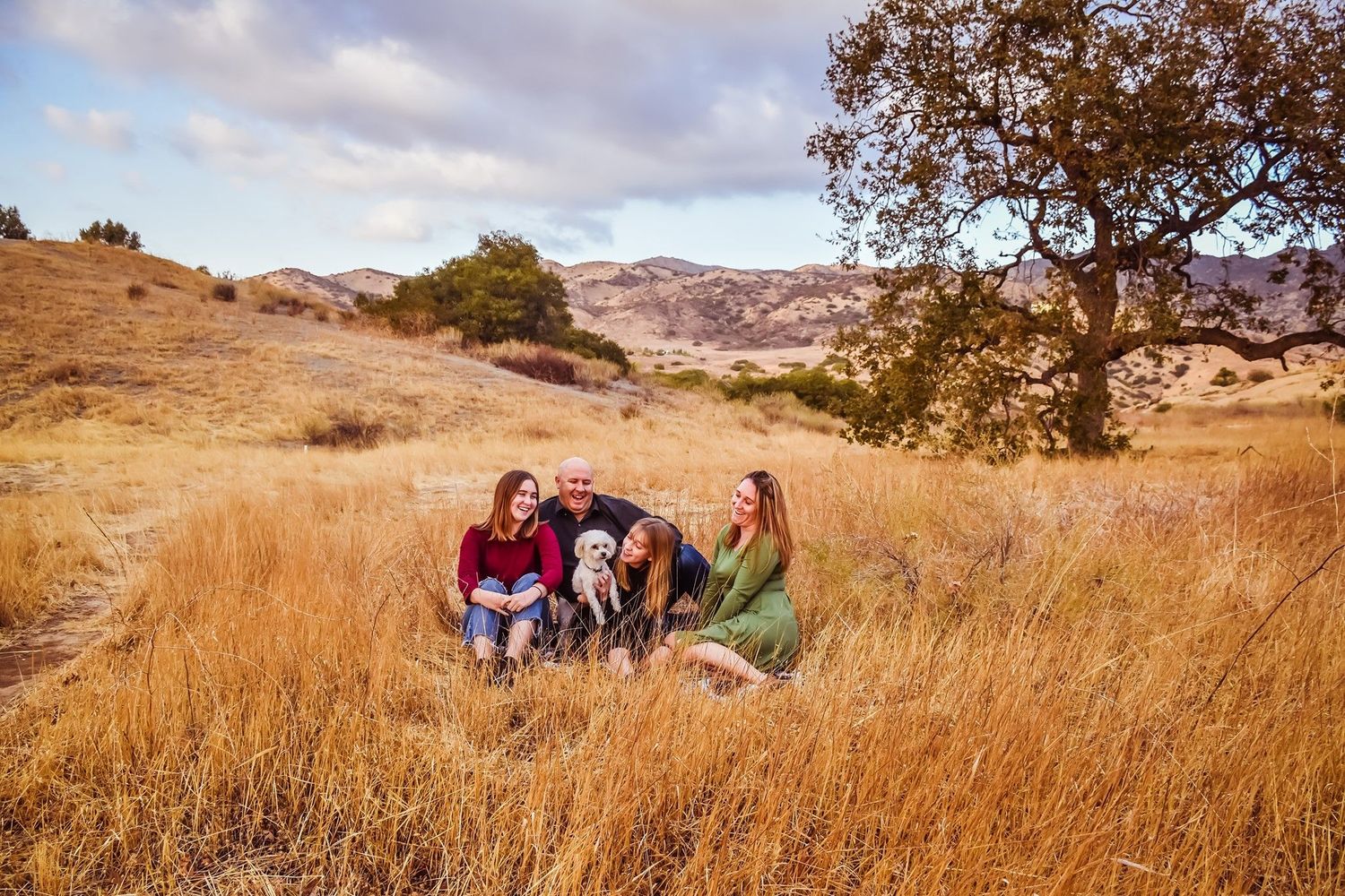 People relax in golden autumn grass on rolling hills with trees and mountains in the background during sunset.