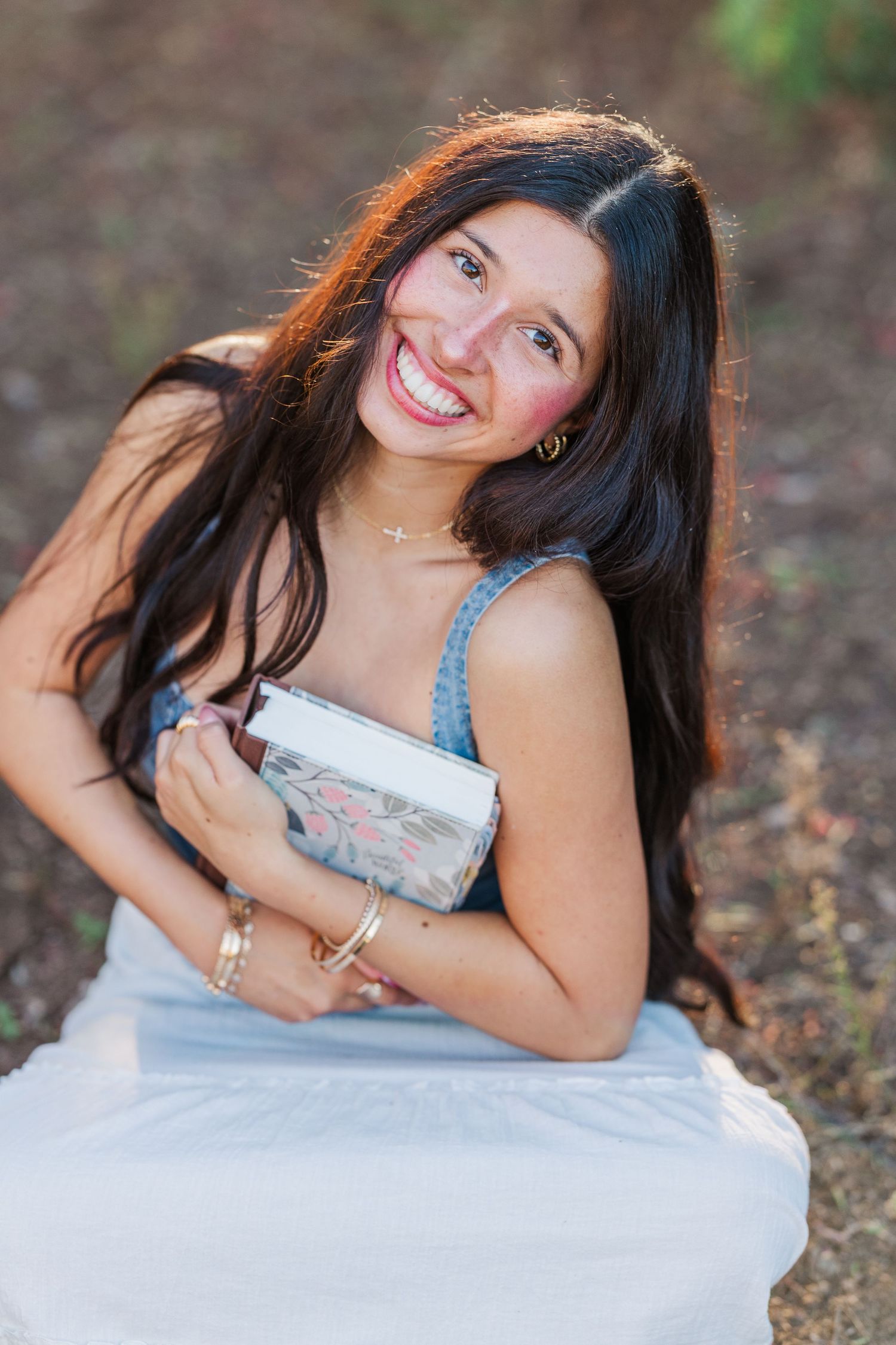 Smiling student in denim top and white skirt holds a book while sitting outdoors during a portrait session.