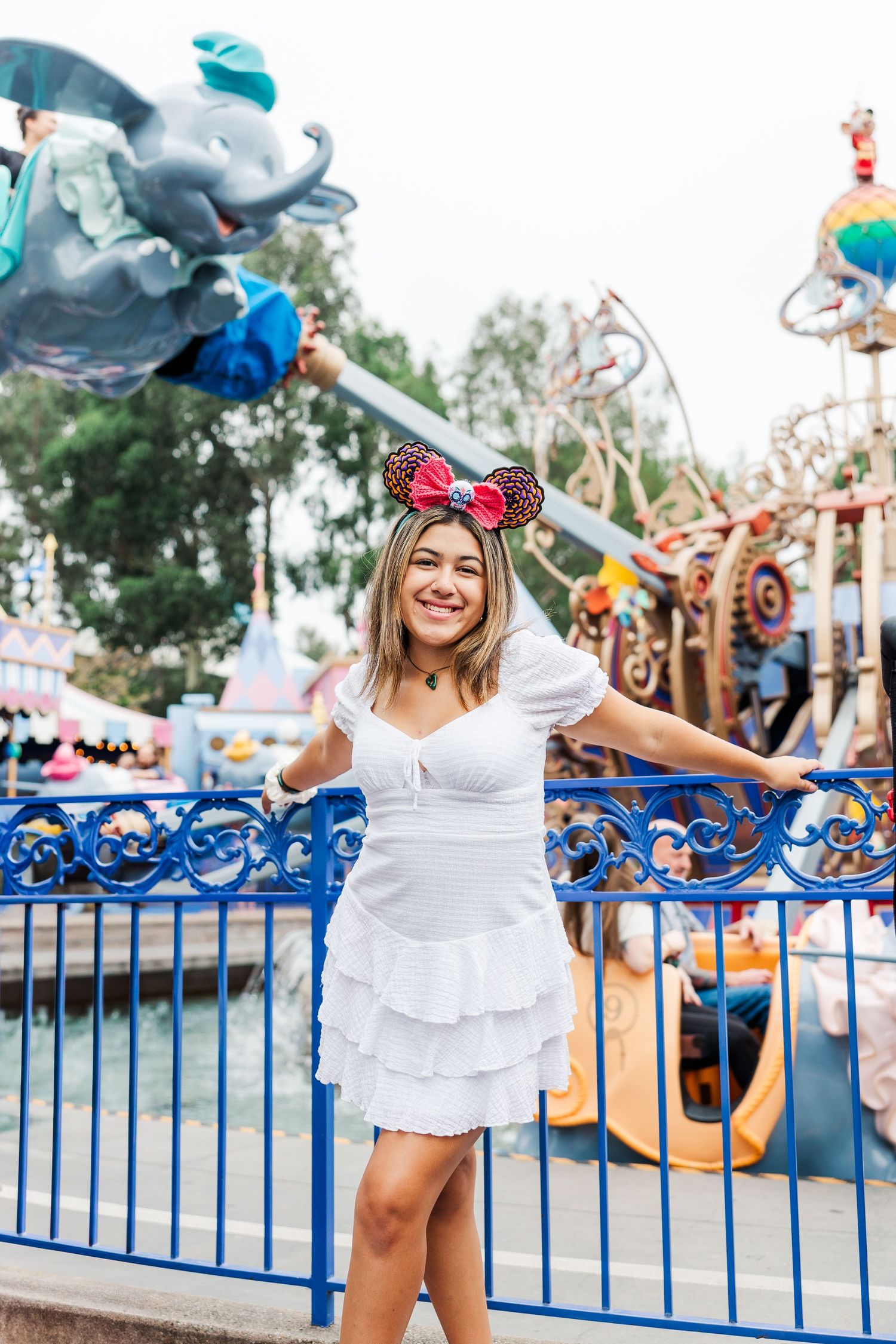 Visitor in white dress and Mickey ears poses joyfully in front of colorful theme park attractions and blue railings.