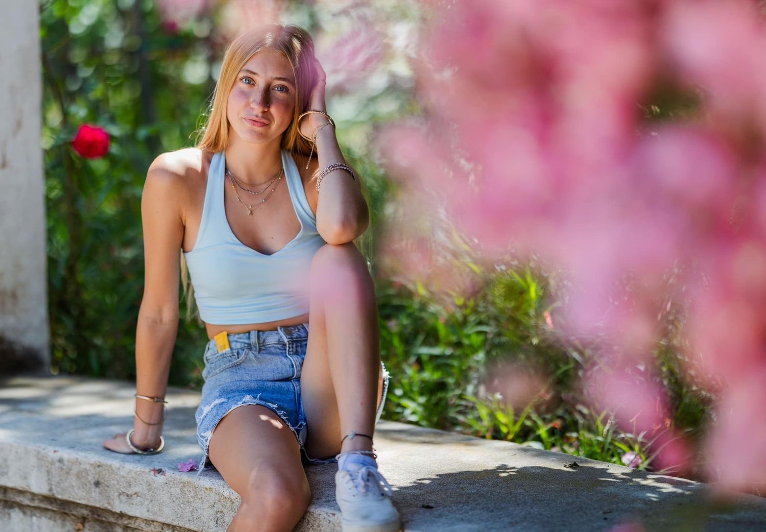 A person in casual summer attire sits on a stone wall with pink flowers in the foreground on a sunny day.