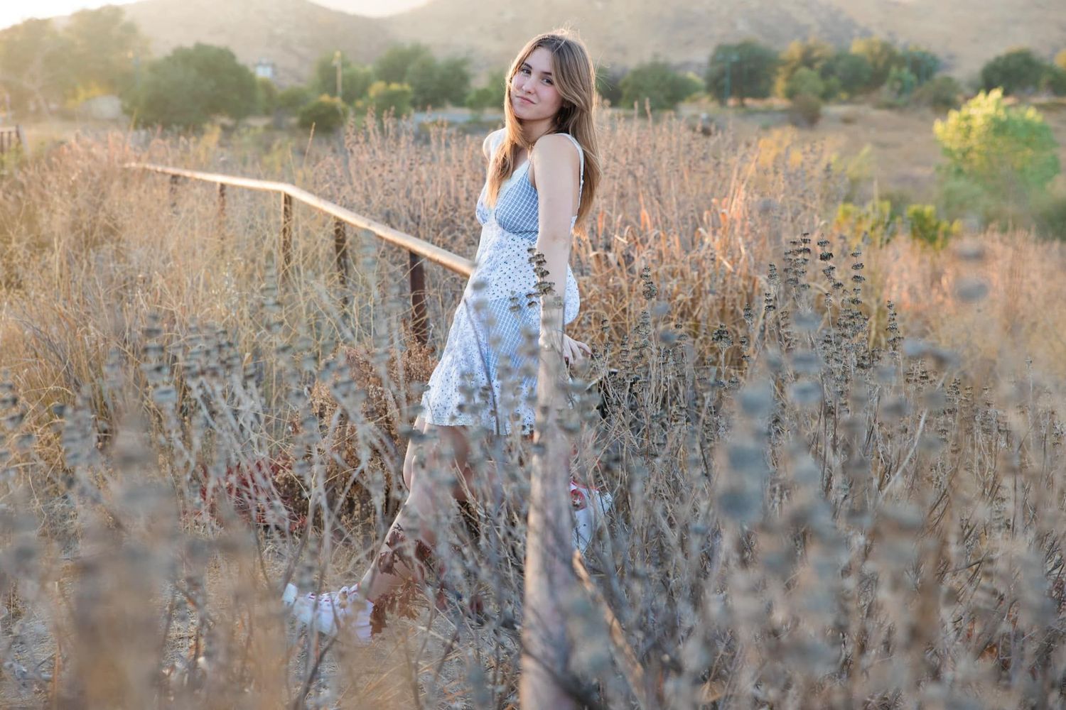 Person in a white sundress standing among tall dry brush in a golden field at sunset.