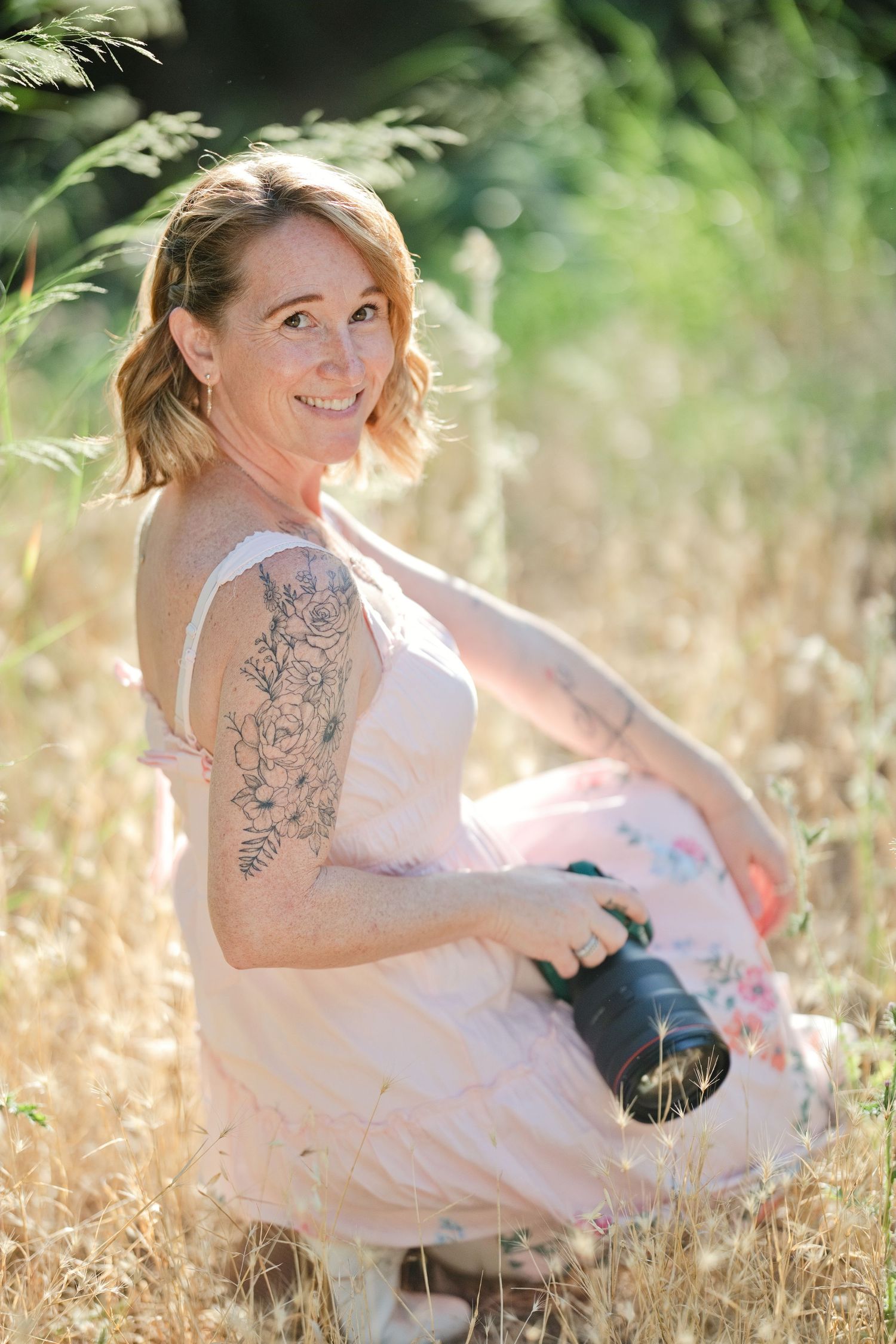 Smiling woman with tattoos in pink floral dress sitting in golden grass field holding a camera on sunny day.