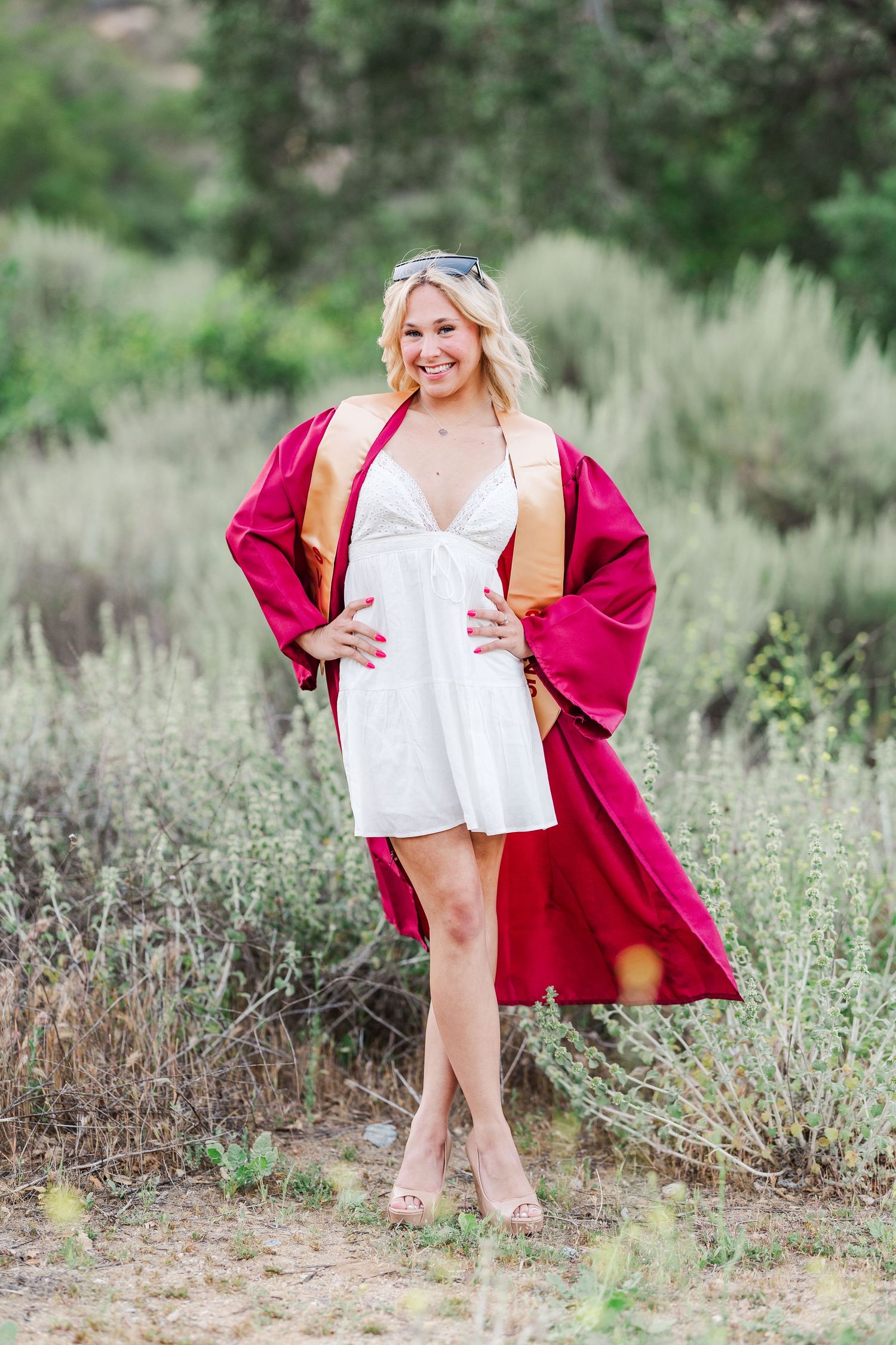 A person in a white dress and burgundy robe poses outdoors among desert sage brush.