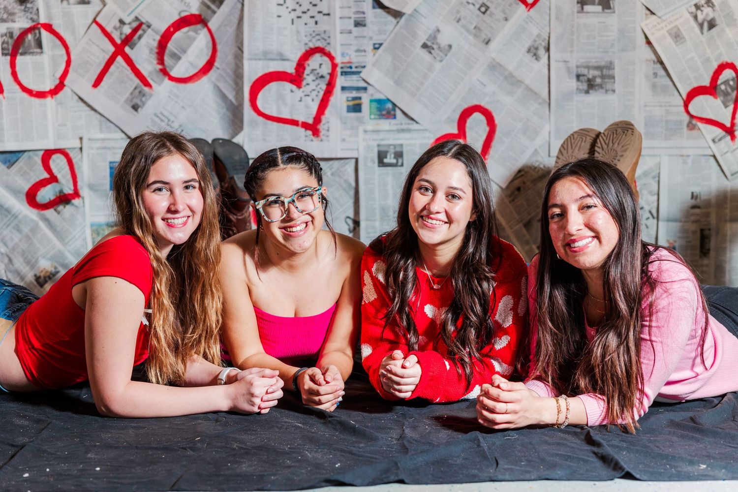 A cheerful group in Valentine's Day themed attire lying on the floor with hearts and xo decorations on the wall behind them.
