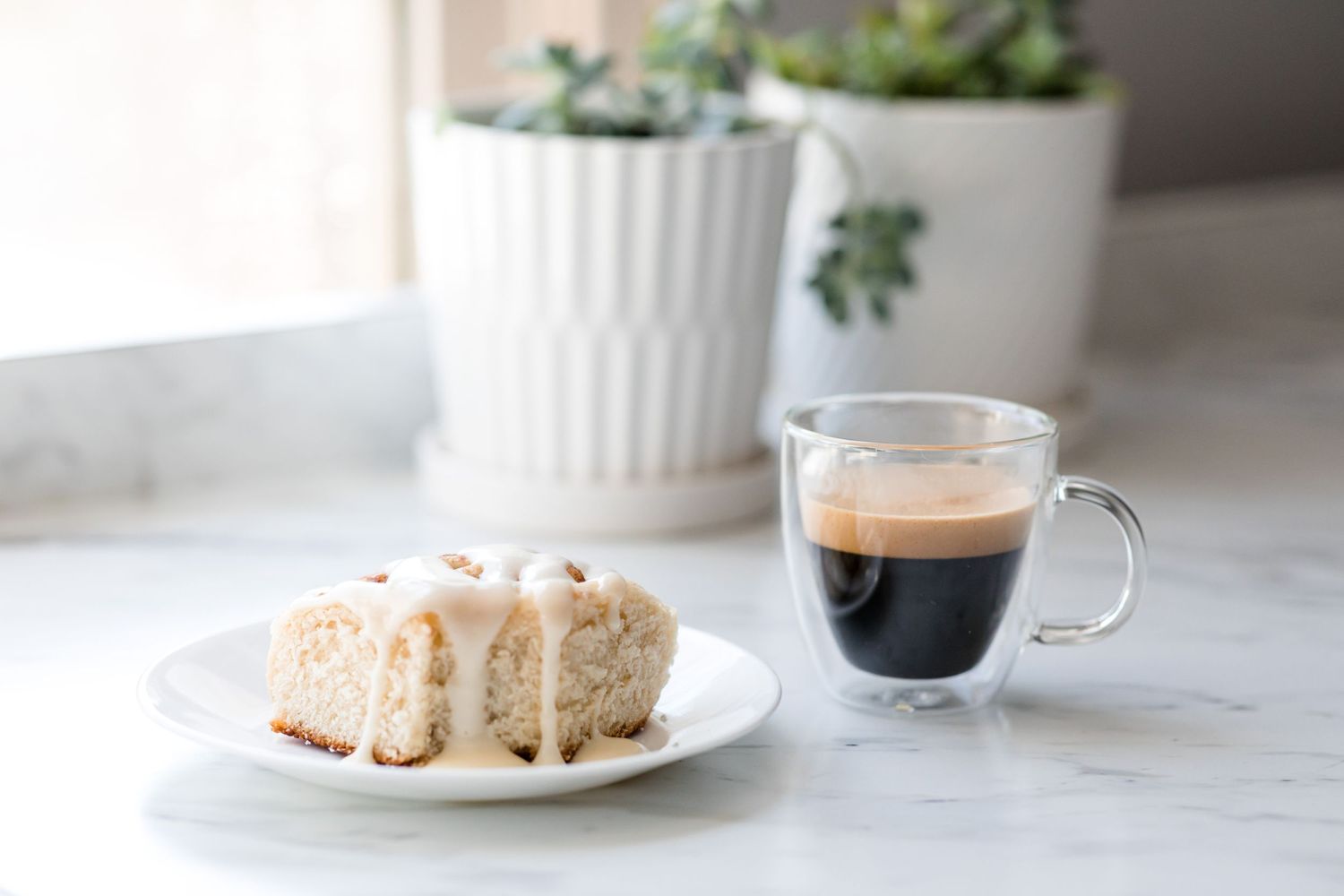A glass of espresso and glazed pastry on a white plate against a bright minimal background.