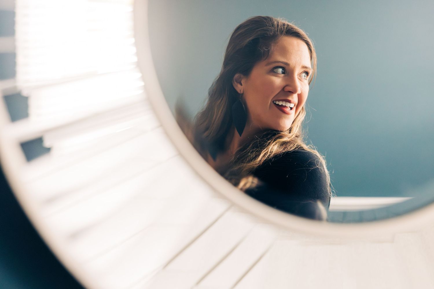 Reflection of woman smiling in round mirror with soft window light.