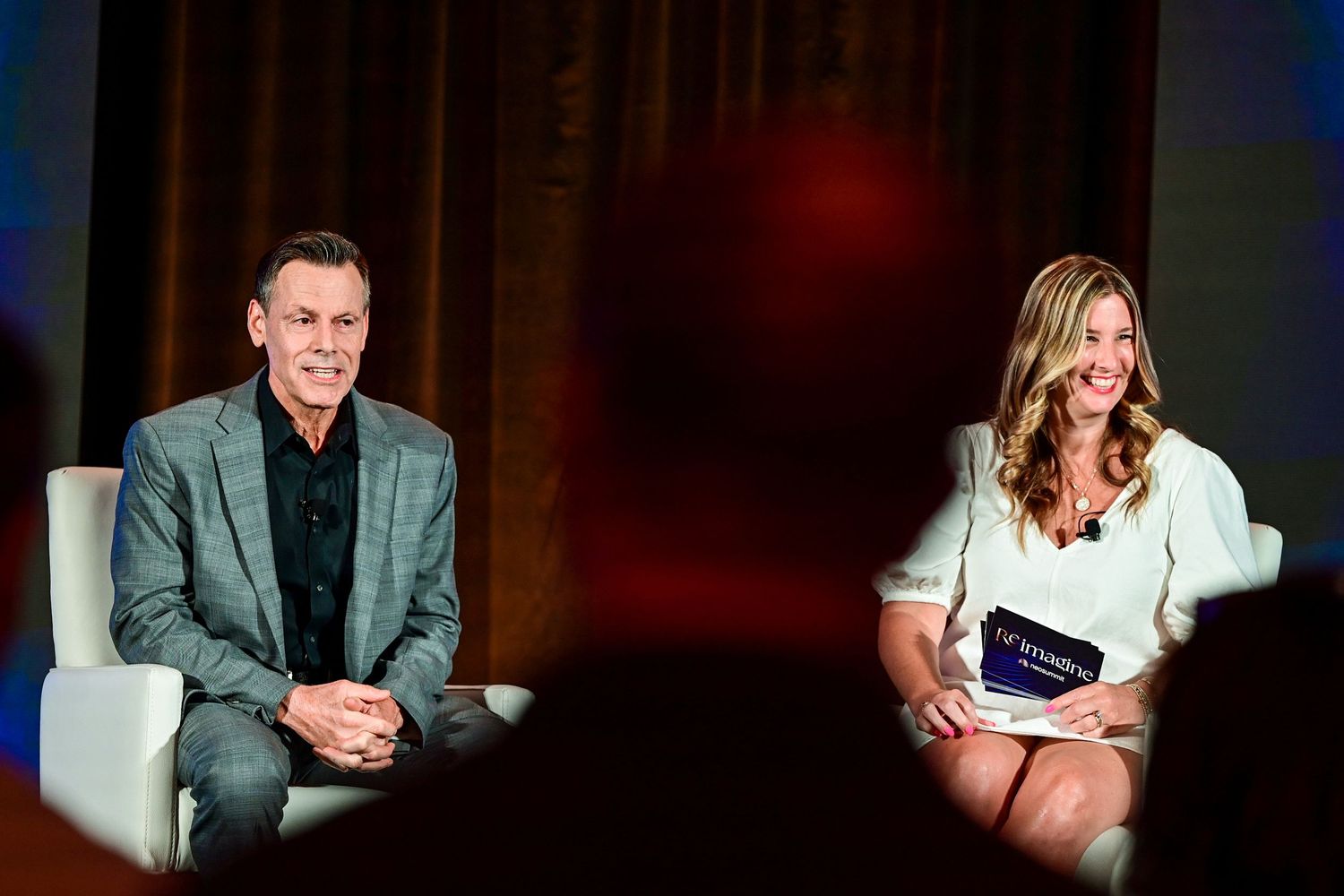 Two speakers, a man in gray suit and woman in white dress, seated on stage during a professional conference panel.