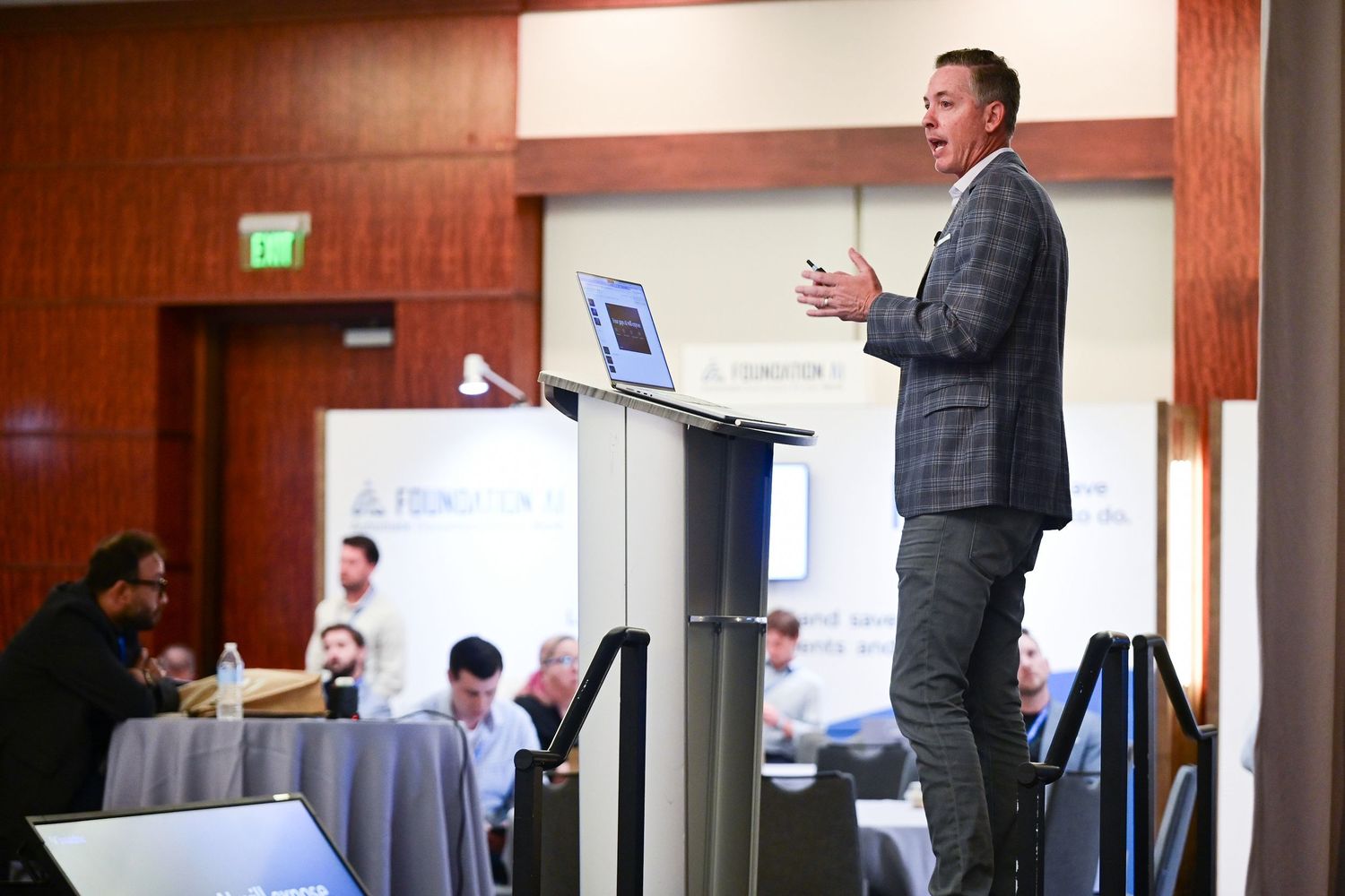 A professional speaker in a plaid suit presents at a podium during a business conference with attendees seated in background.