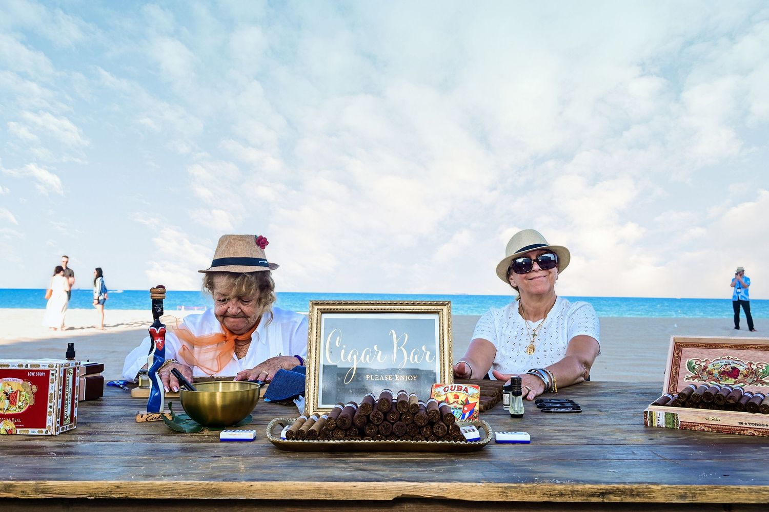 Two people in hats sitting behind a cigar bar table on a scenic beach setting.