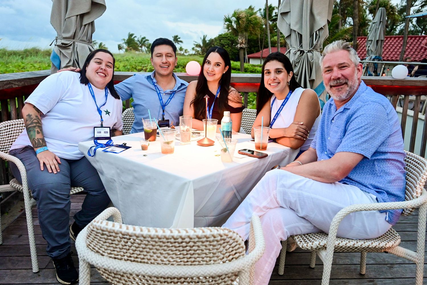 Five people smile together at an outdoor dining table with drinks and desserts at a waterfront restaurant.