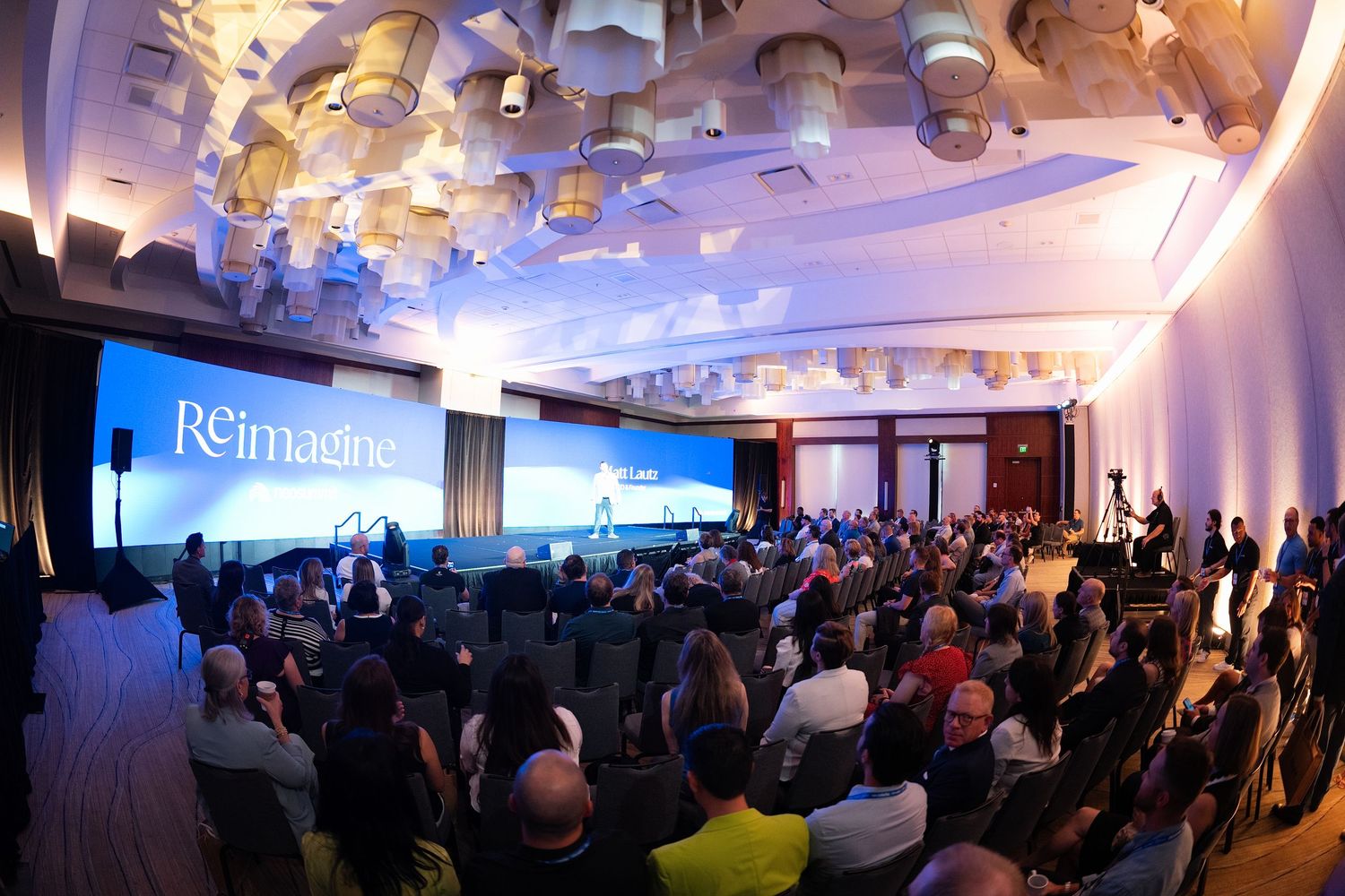 Large conference audience gathered at a Reimagine event with a blue presentation screen on stage.